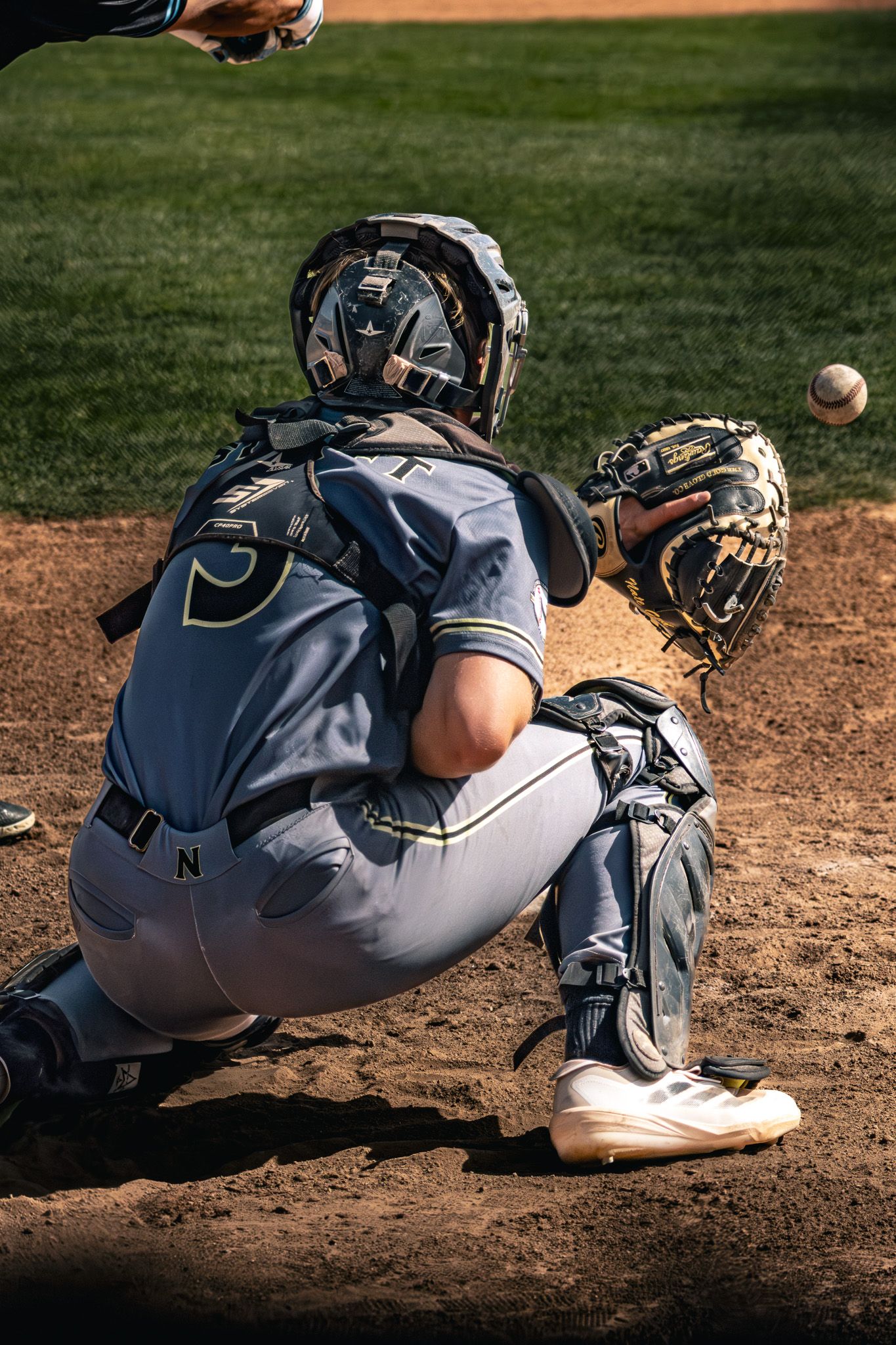 Baseball catcher in full gear crouches to receive incoming pitch.