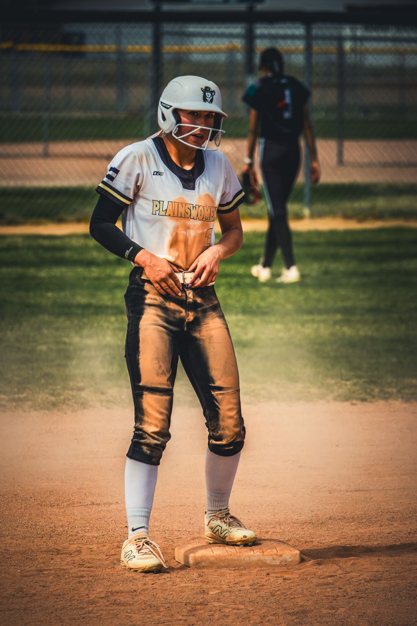 Dirt-covered softball player stands on base in Plainsmen uniform during sunny game.