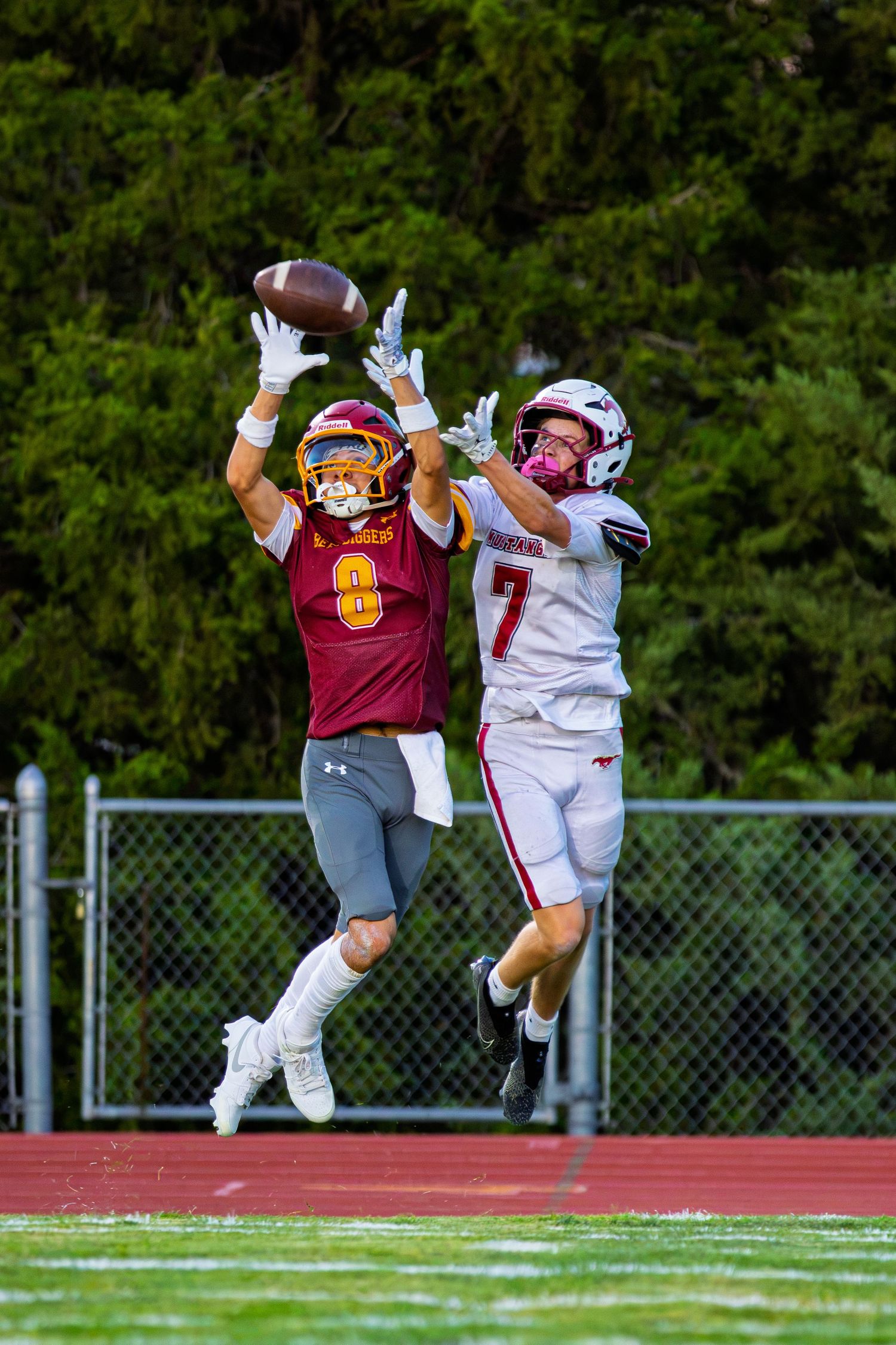 Two football players jump for a pass, one in maroon #8 and one in white #7, reaching for the ball.