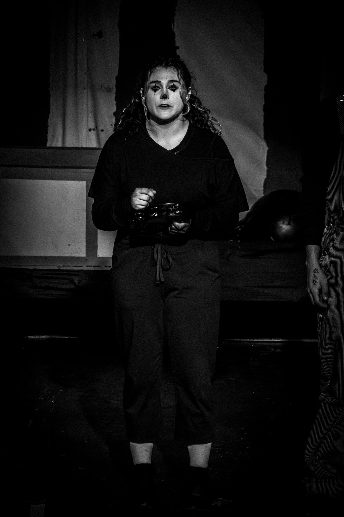 A performer in black costume with dramatic clown makeup stands on stage in a black and white theatrical production.