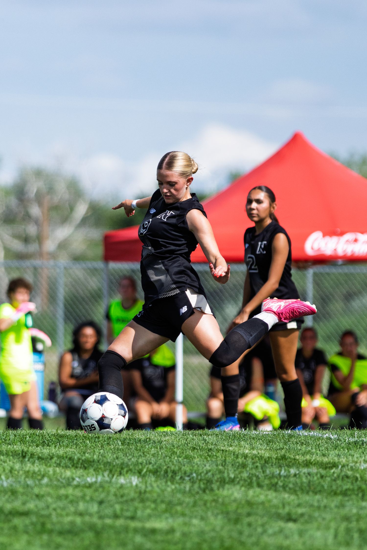 Young female soccer player in black uniform kicking ball during outdoor match with Coca-Cola tent in background.