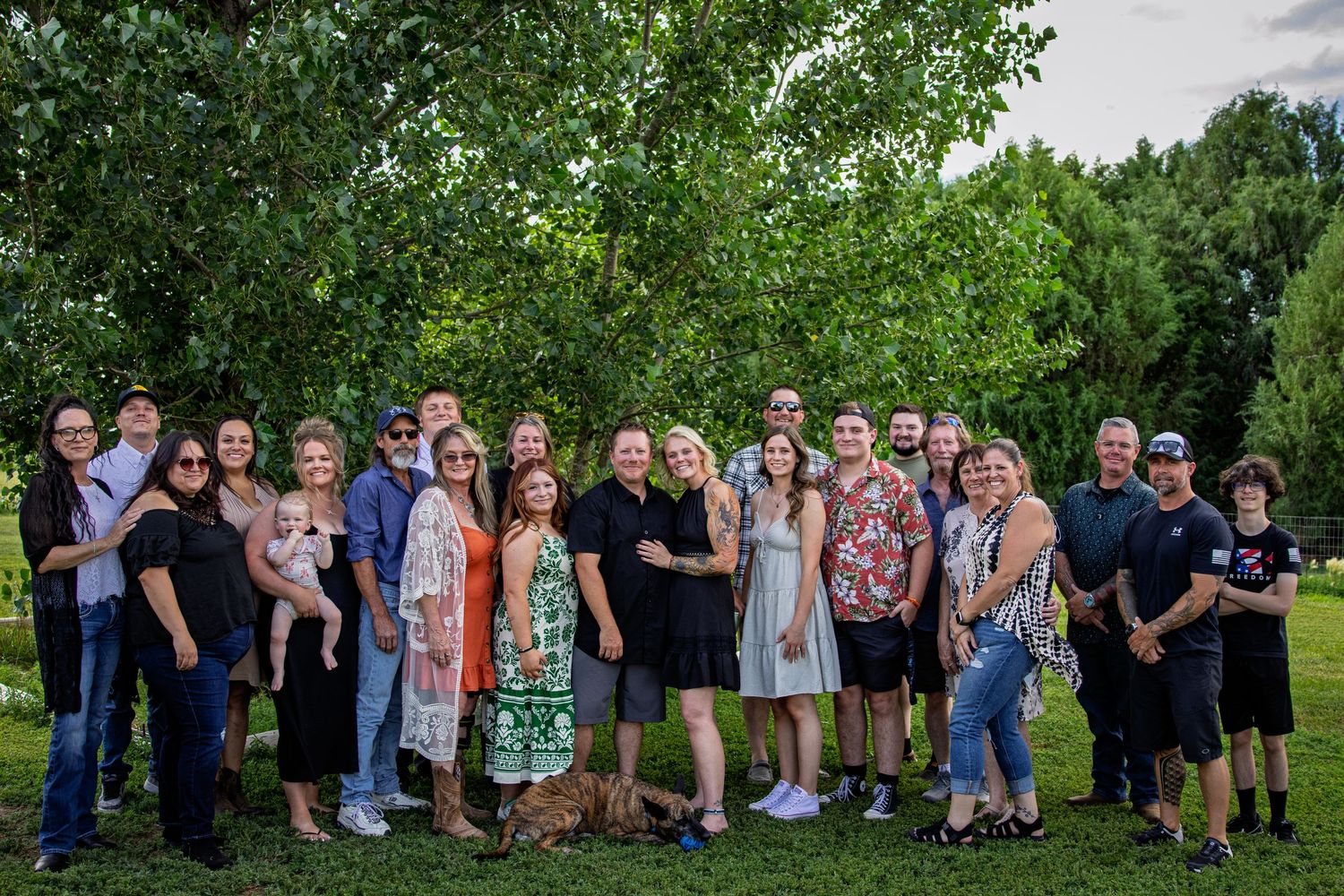 Large extended family group smiling together outdoors on green grass with lush trees behind.