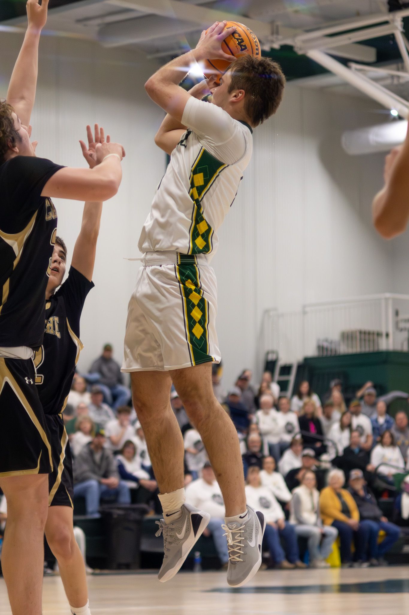 A high school basketball player in white and green uniform jumps to shoot over defenders during an indoor game.