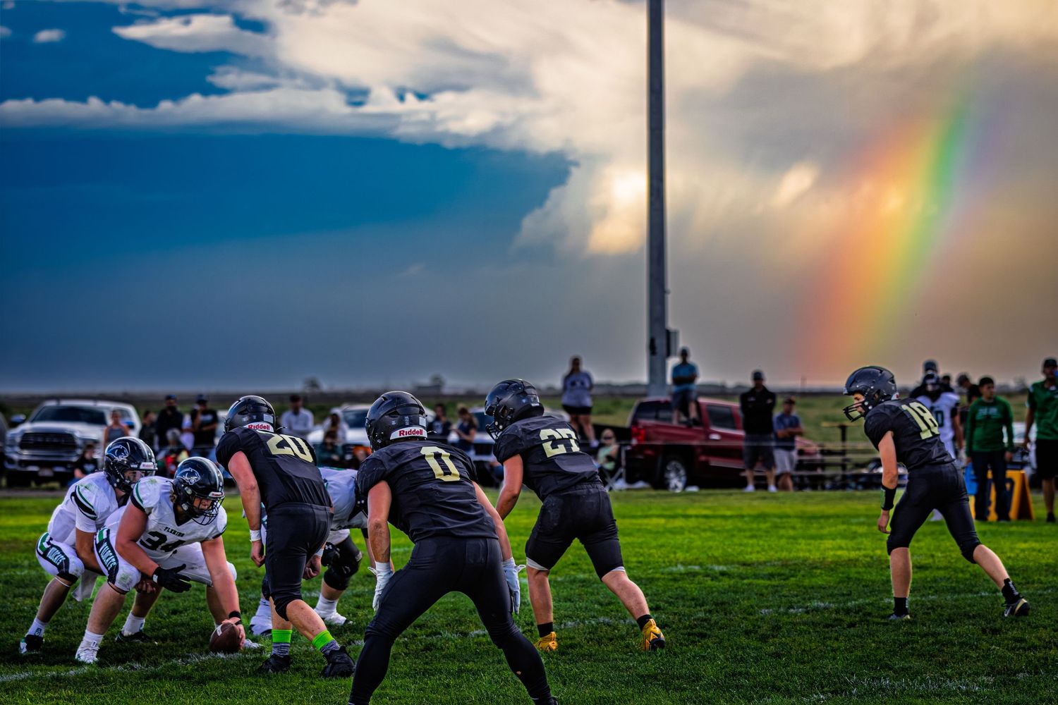Football players line up for a play on a stormy evening with a rainbow visible in the background.