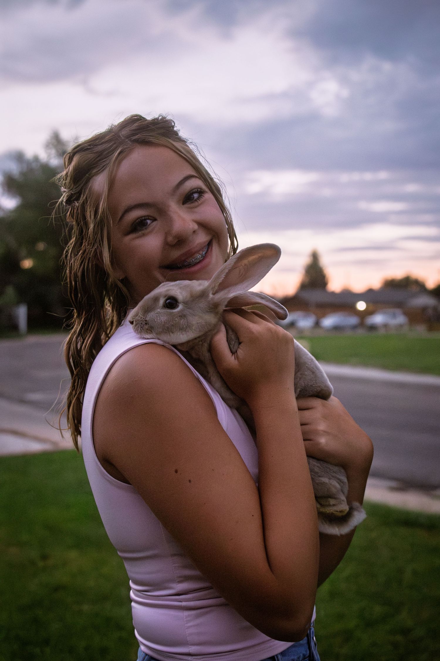 Smiling girl with braces holding a gray rabbit close to her face at sunset outdoors.