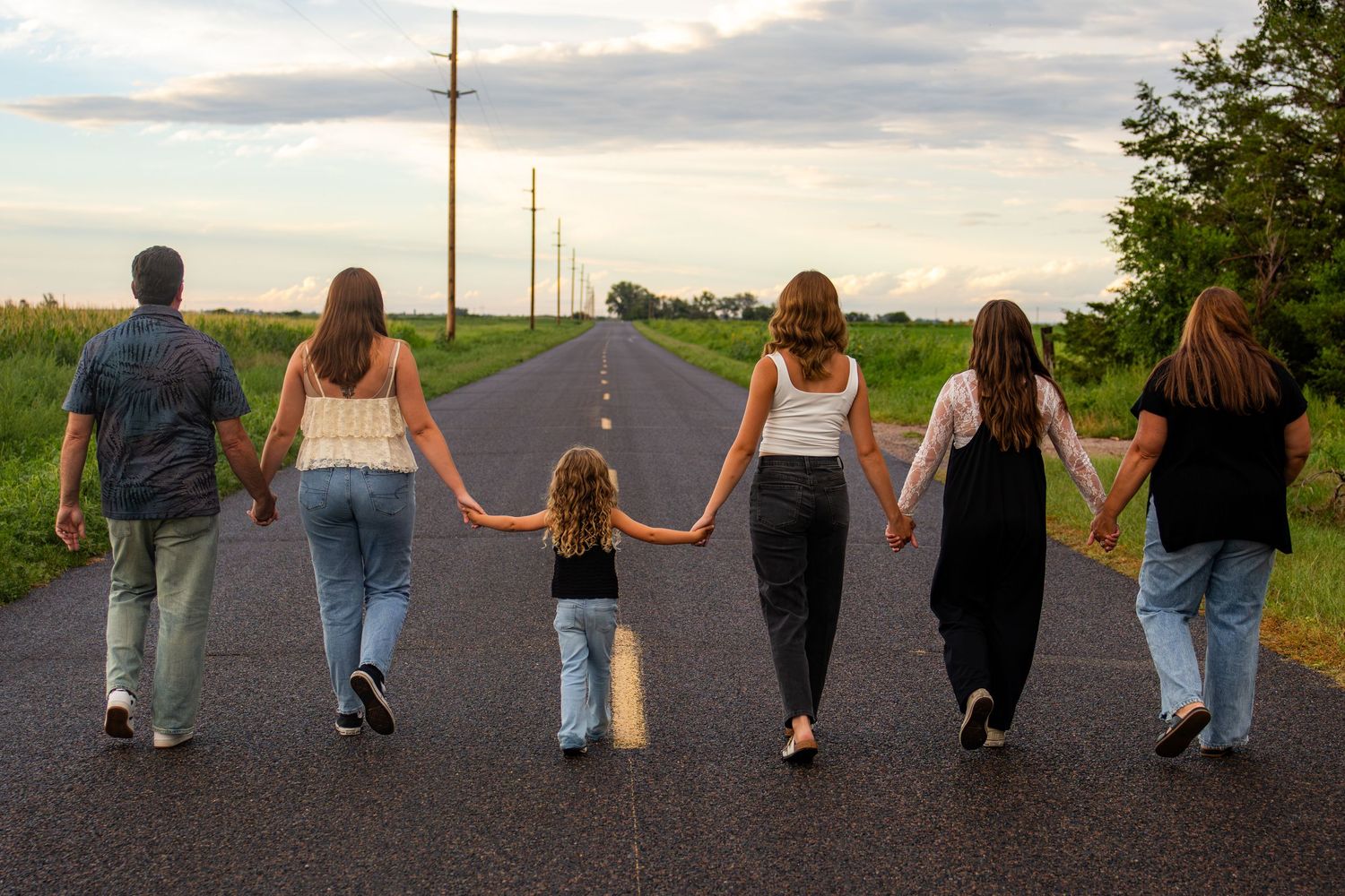 Family of six walking hand-in-hand down a straight rural road at dusk, viewed from behind.