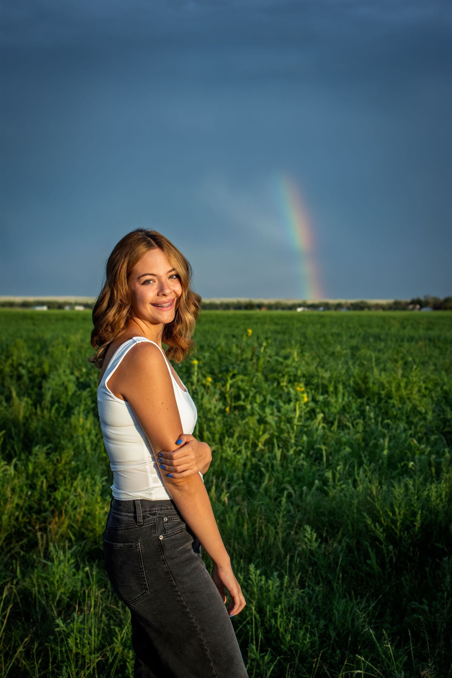 Young woman in white tank top and gray jeans posing in green field with rainbow in cloudy sky.