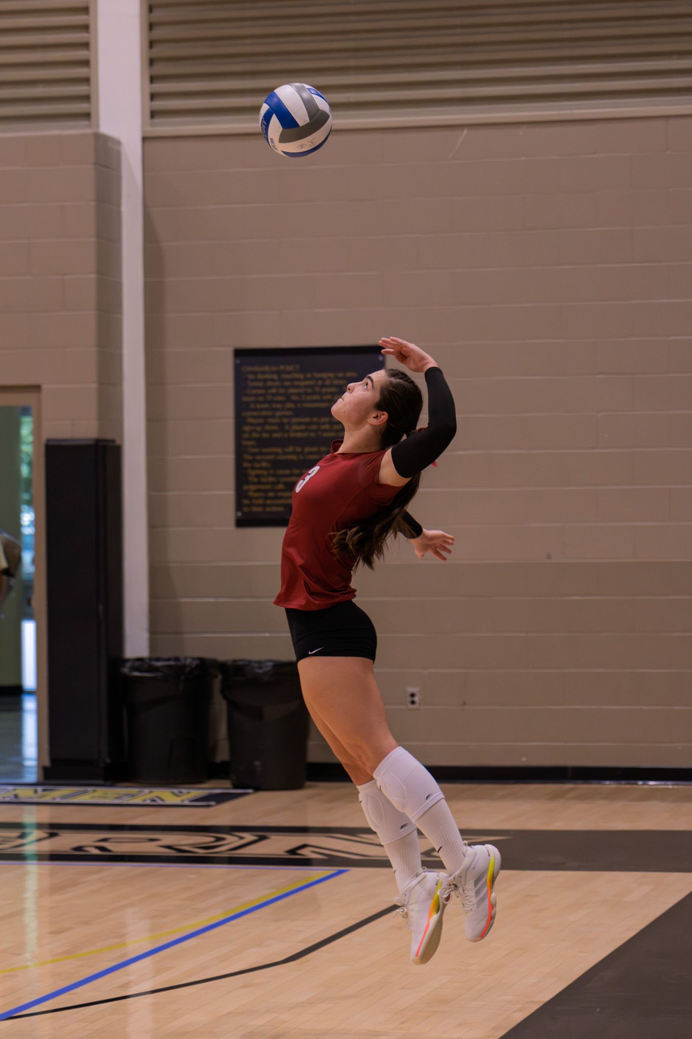 Female volleyball player in red jersey jumping to serve the ball in an indoor gymnasium.