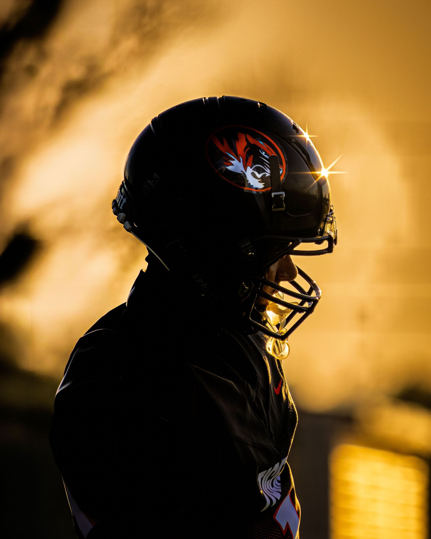Dramatic silhouette of a Tigers football player wearing a helmet against a golden sunset sky.
