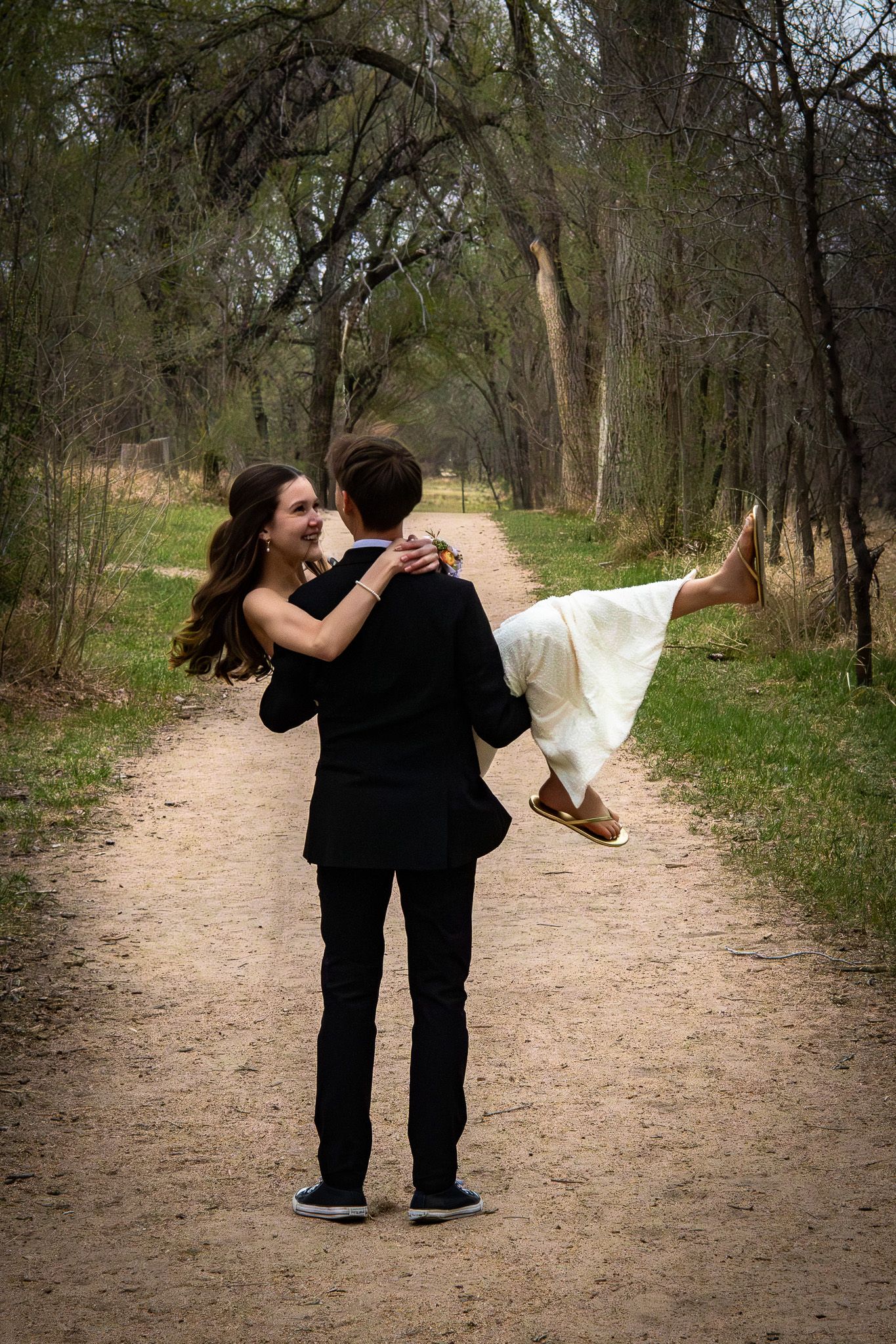 Man in black suit carrying a woman in a white dress down a wooded forest path.