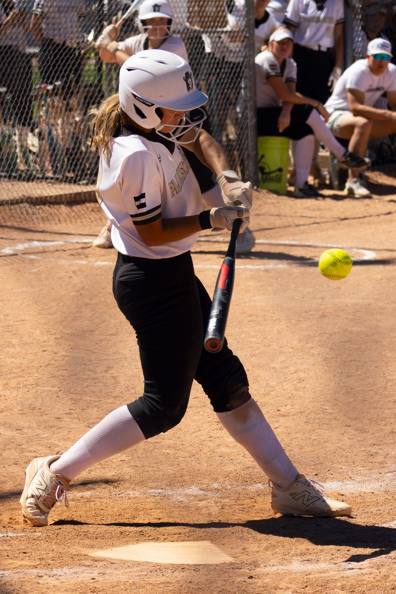Female softball player in white uniform swinging bat at yellow ball during game.