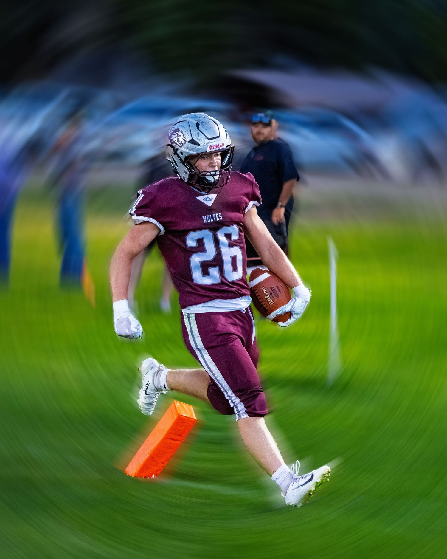 Football player number 26 in maroon uniform running with ball near orange pylon with radial blur effect.