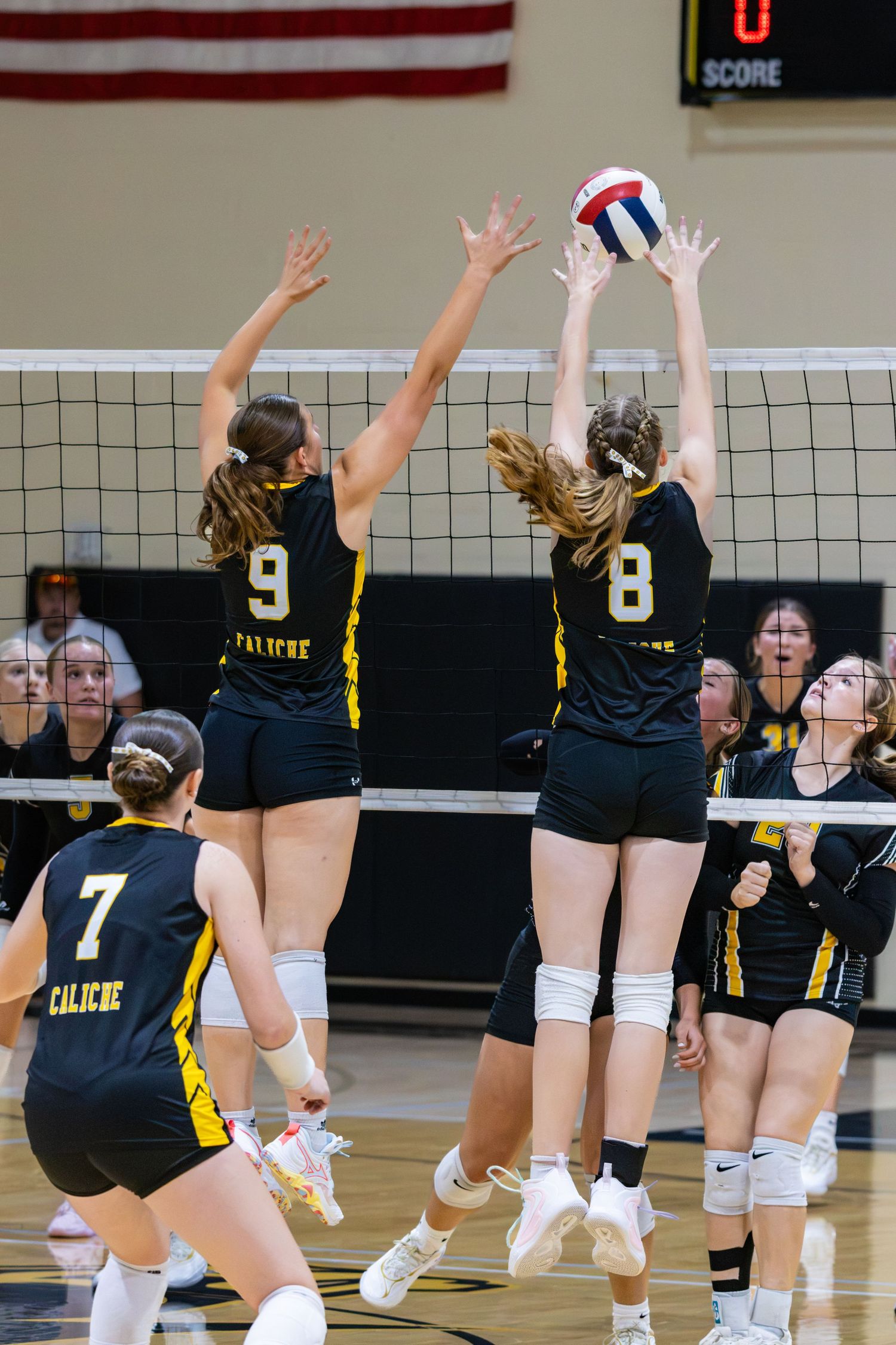 Caliche volleyball players numbers 9 and 8 jump to block ball at net during game.