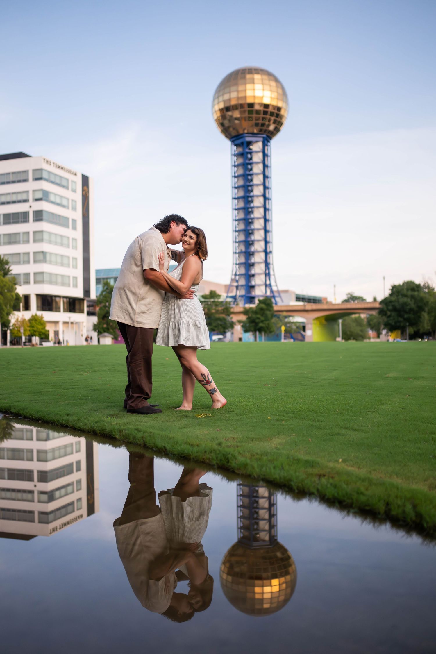 A couple shares a kiss on a green lawn with the Sunsphere tower reflected in a nearby pond in Knoxville.