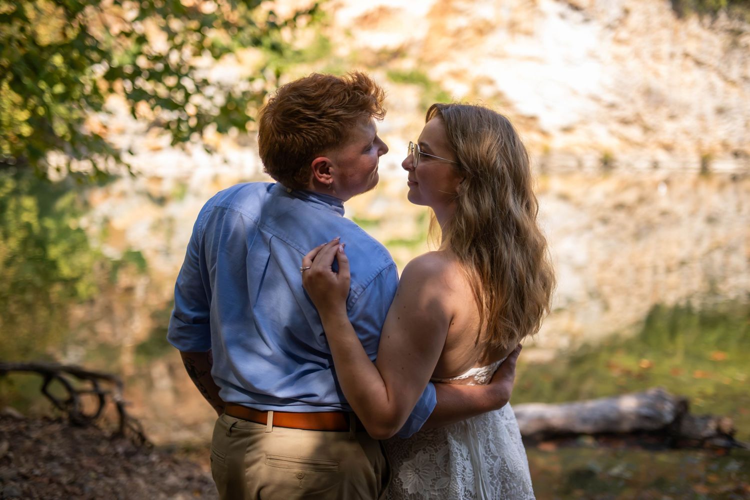 A couple shares a romantic moment outdoors near a rocky waterfall, the man in blue shirt and woman in white dress.