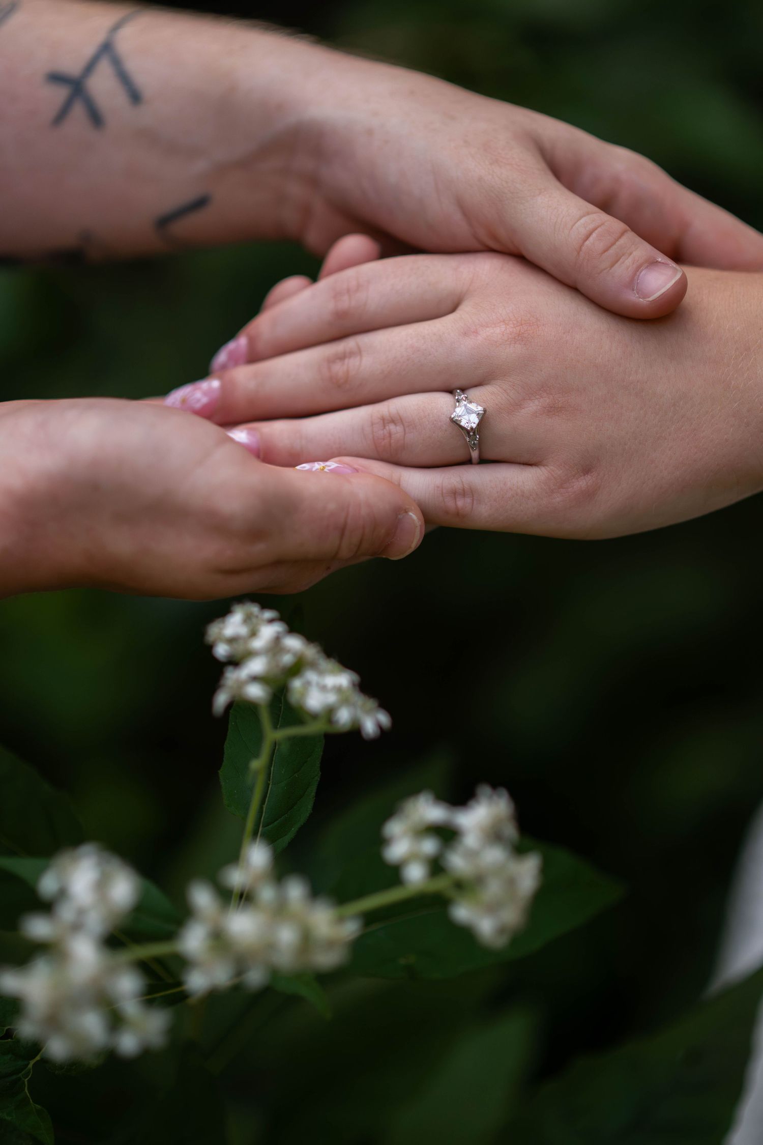 Close-up of hands holding, featuring a diamond engagement ring, with white flowers in the background.