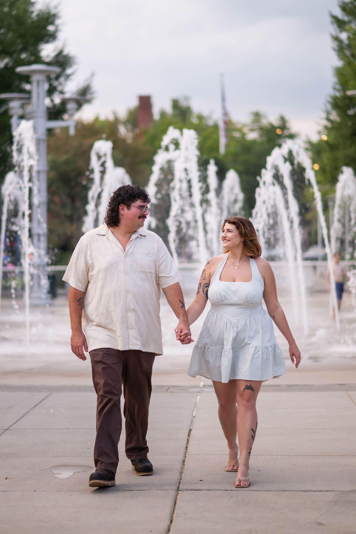 A smiling couple holds hands while walking past fountains in an outdoor plaza at dusk.
