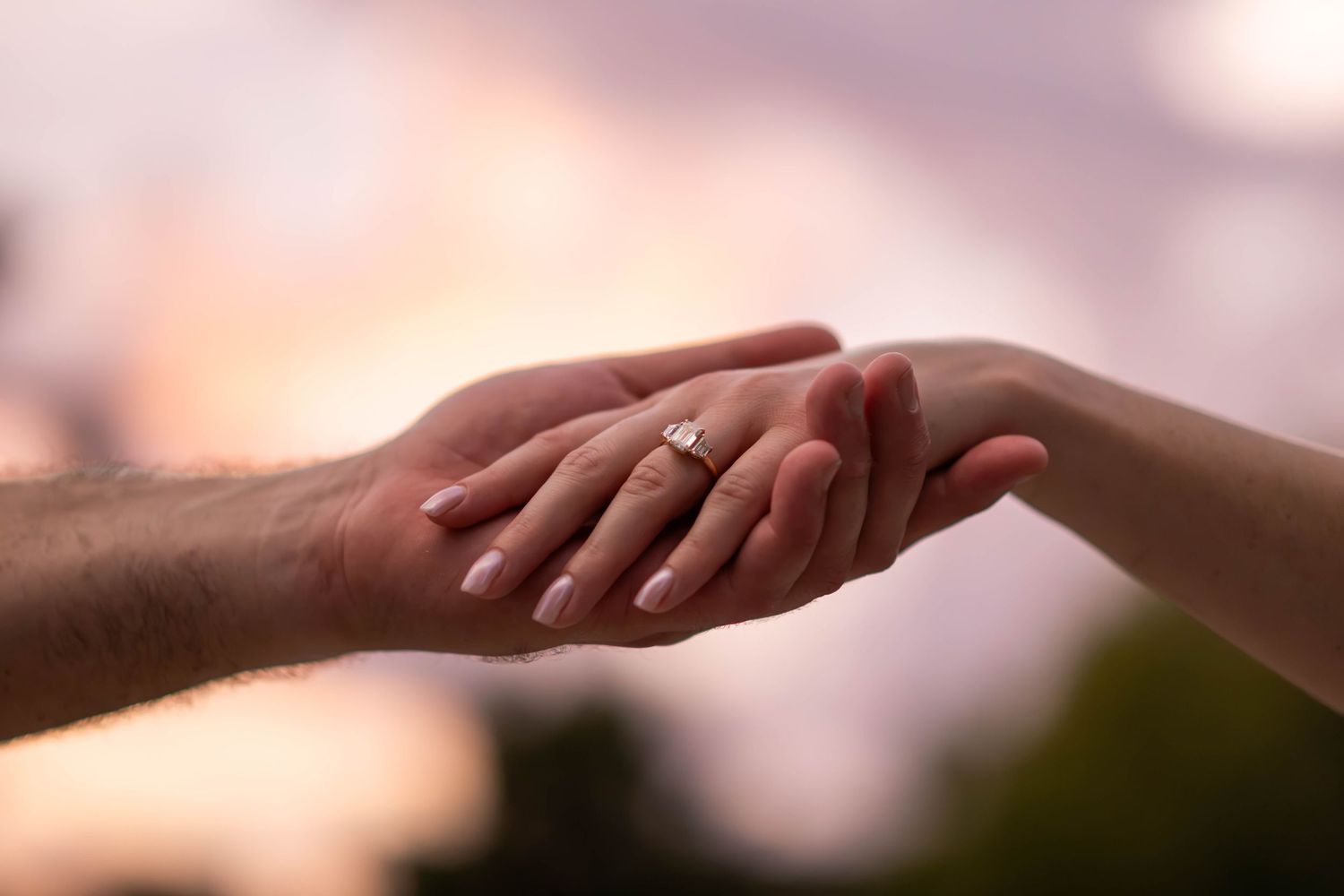 Two hands gently touch as one displays a diamond engagement ring against a soft pink sunset sky.
