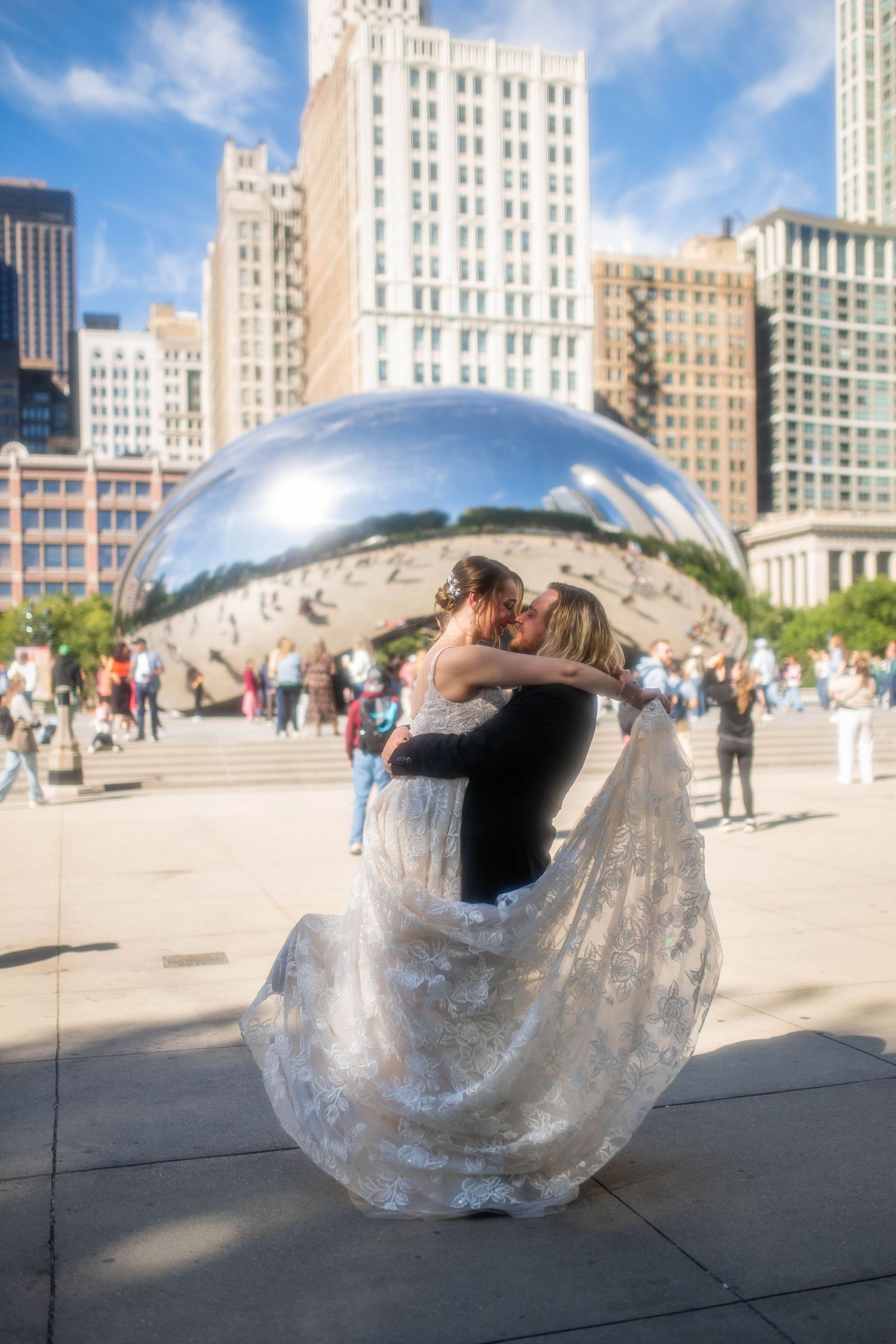 Bride in a lace gown and groom in a dark suit embrace and kiss in front of Cloud Gate sculpture in Chicago's Millennium Park.