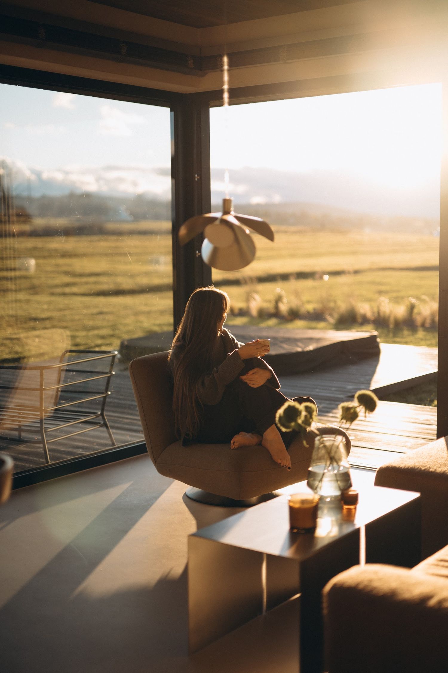 Woman sitting in a modern chair enjoying golden sunset views over a rural landscape through floor-to-ceiling windows.