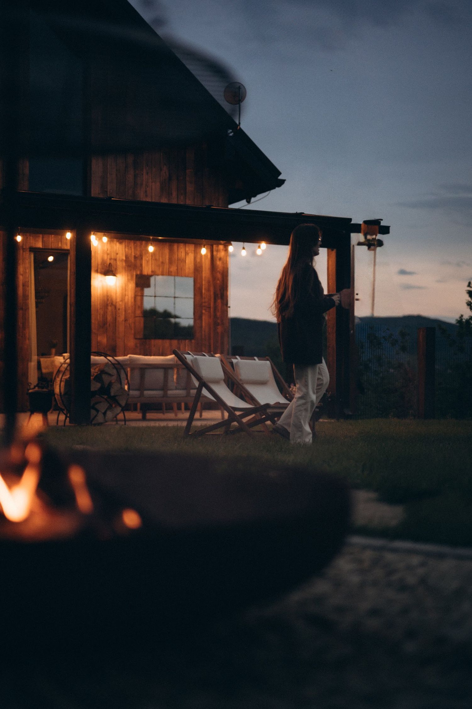 Woman walking toward a warmly lit wooden cabin at night with a fire pit glowing in the foreground.