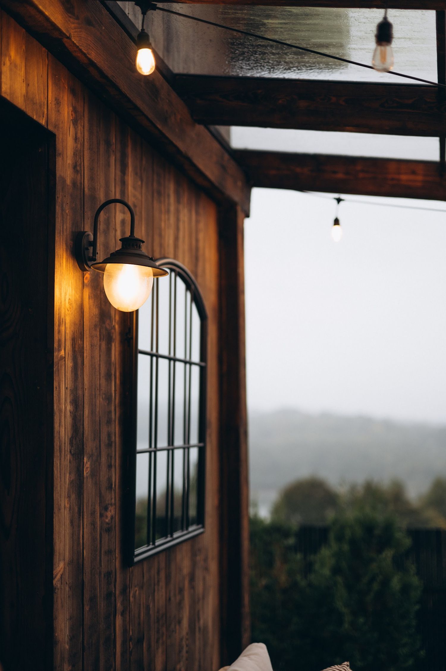 Rustic wooden cabin exterior at dusk with a glowing wall lantern and string lights in rain.