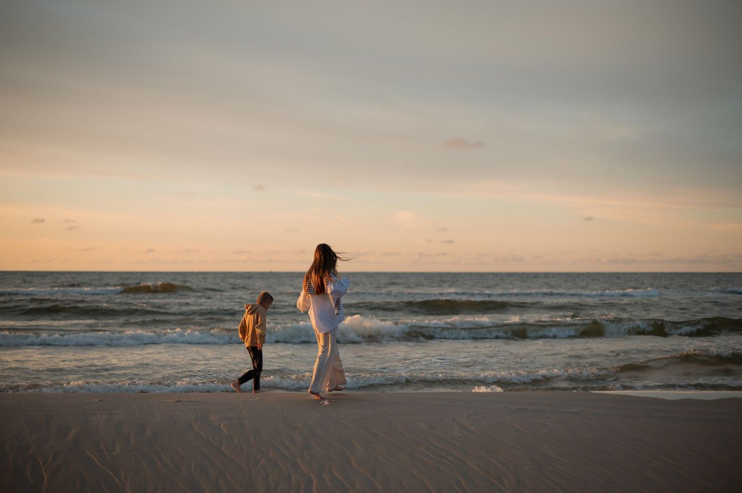 Mother and child walking along a sandy beach at sunset with gentle waves in the background.