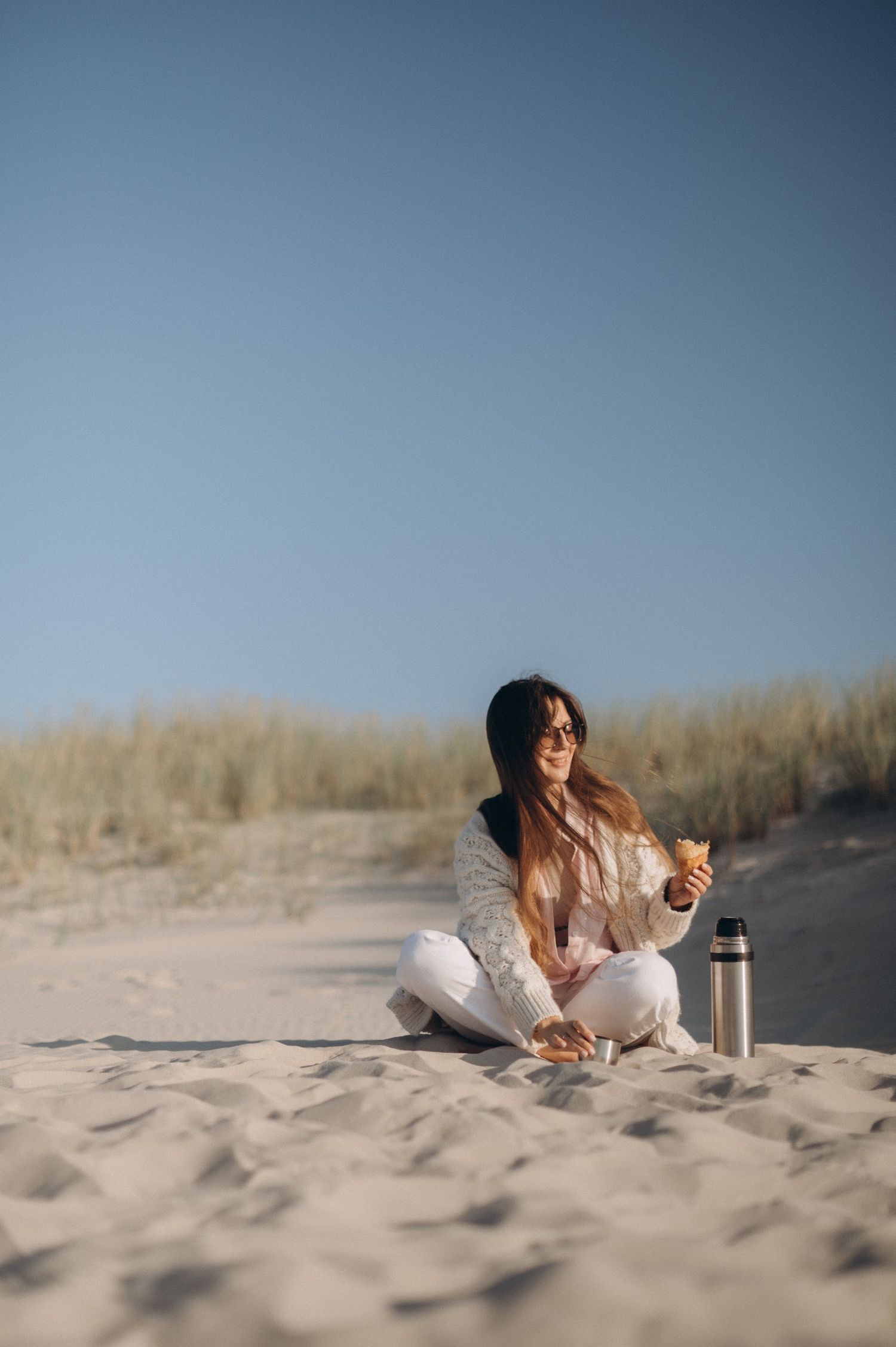 Woman sitting cross-legged on sandy beach dunes, smiling and holding a snack with a thermos beside her.