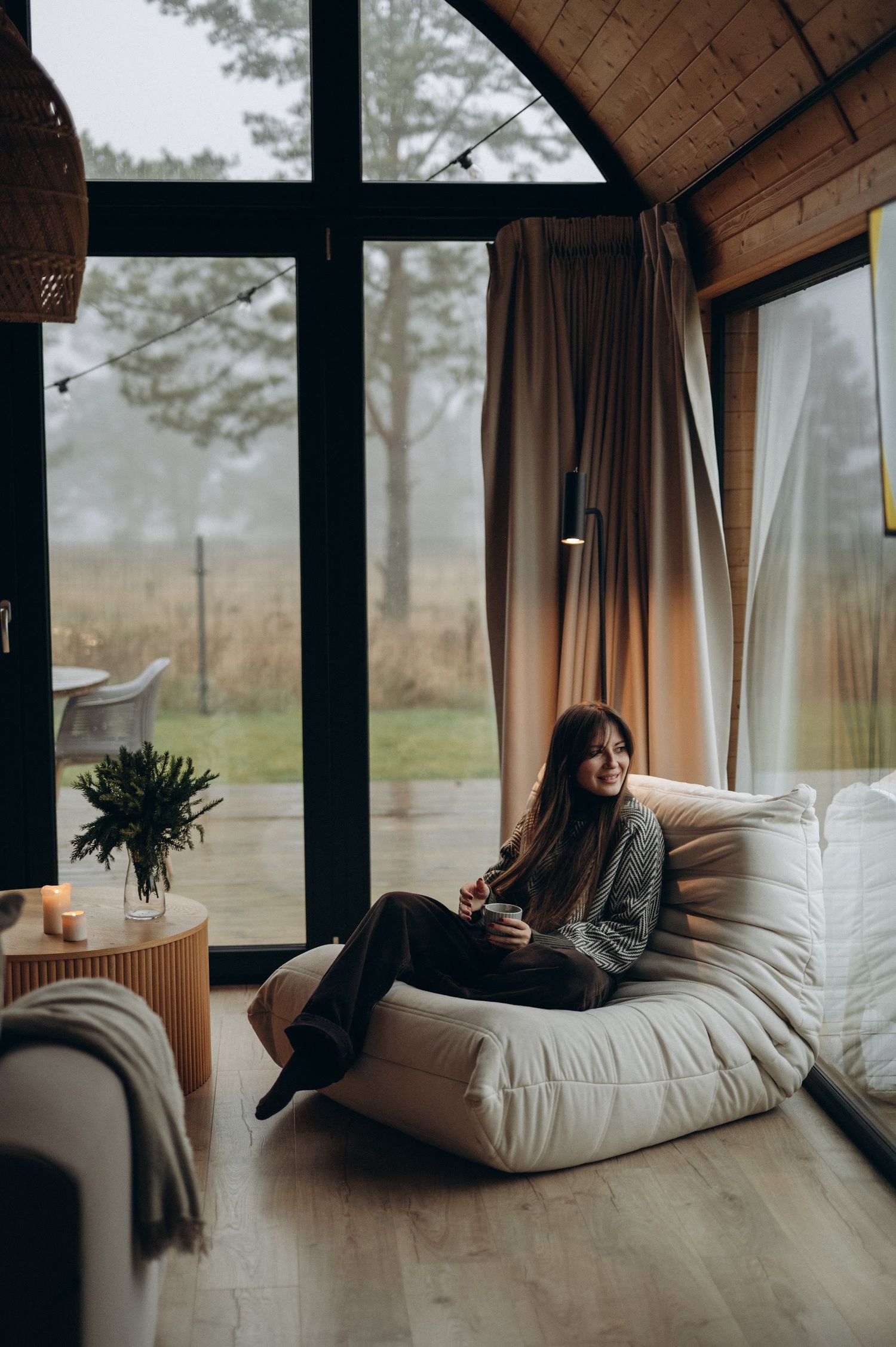 Woman relaxing on a white bean bag chair inside a cozy modern cabin with large windows.
