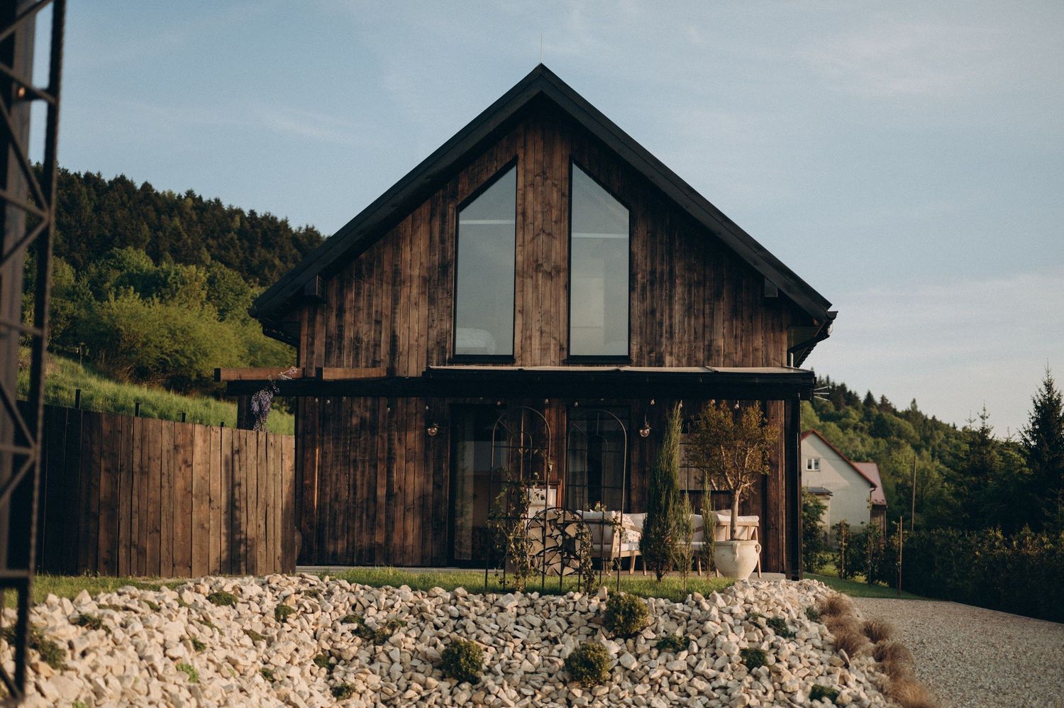 Rustic wooden barn-style house with vertical timber cladding and large gable window set against forested hills.