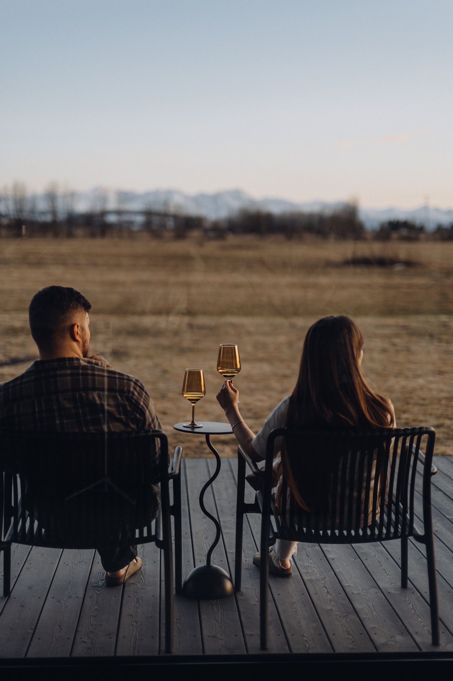 Couple sitting on a wooden deck at dusk, raising wine glasses while overlooking a scenic mountain landscape.