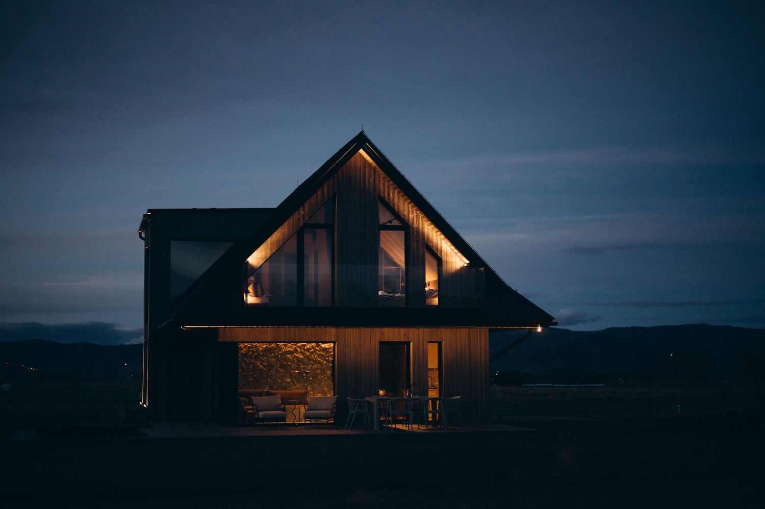 Modern A-frame house illuminated with warm lights against a dramatic dark twilight sky.