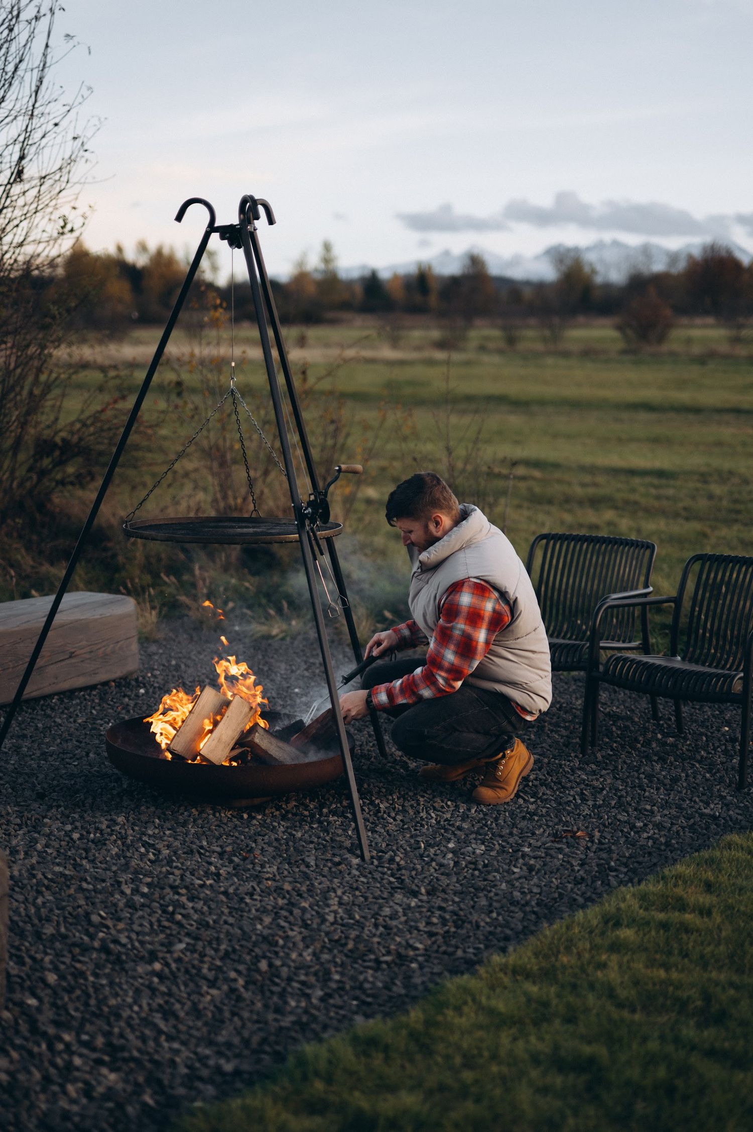 Man tending to outdoor fire pit with cooking tripod at dusk in a scenic countryside setting.