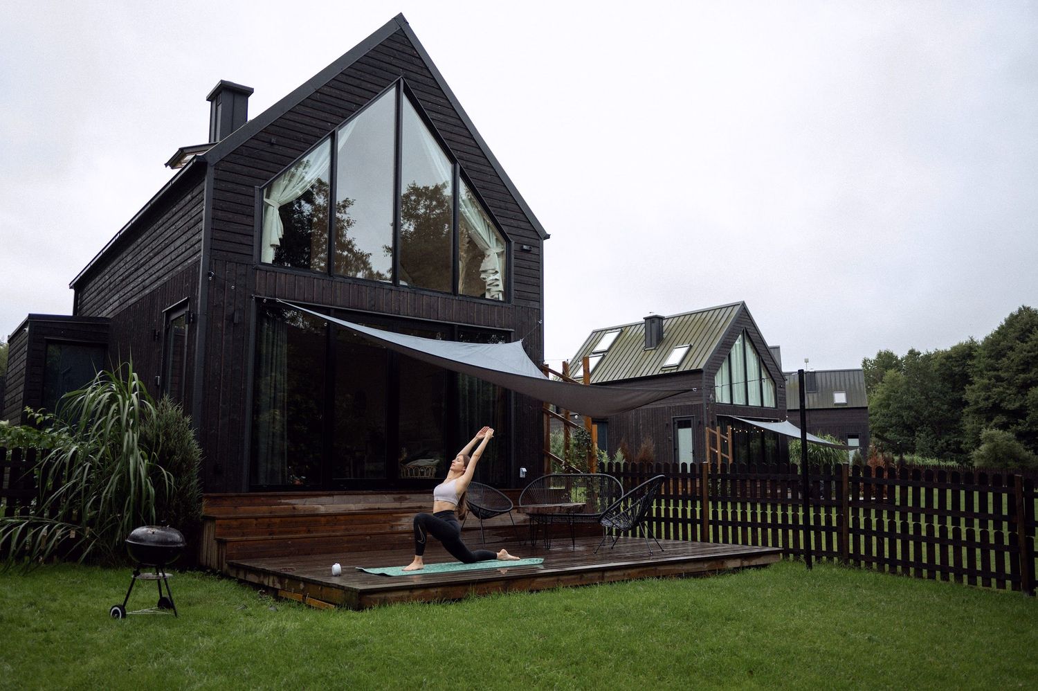 Woman practicing yoga on wooden deck outside a dark modern barn-style house with large glass windows.