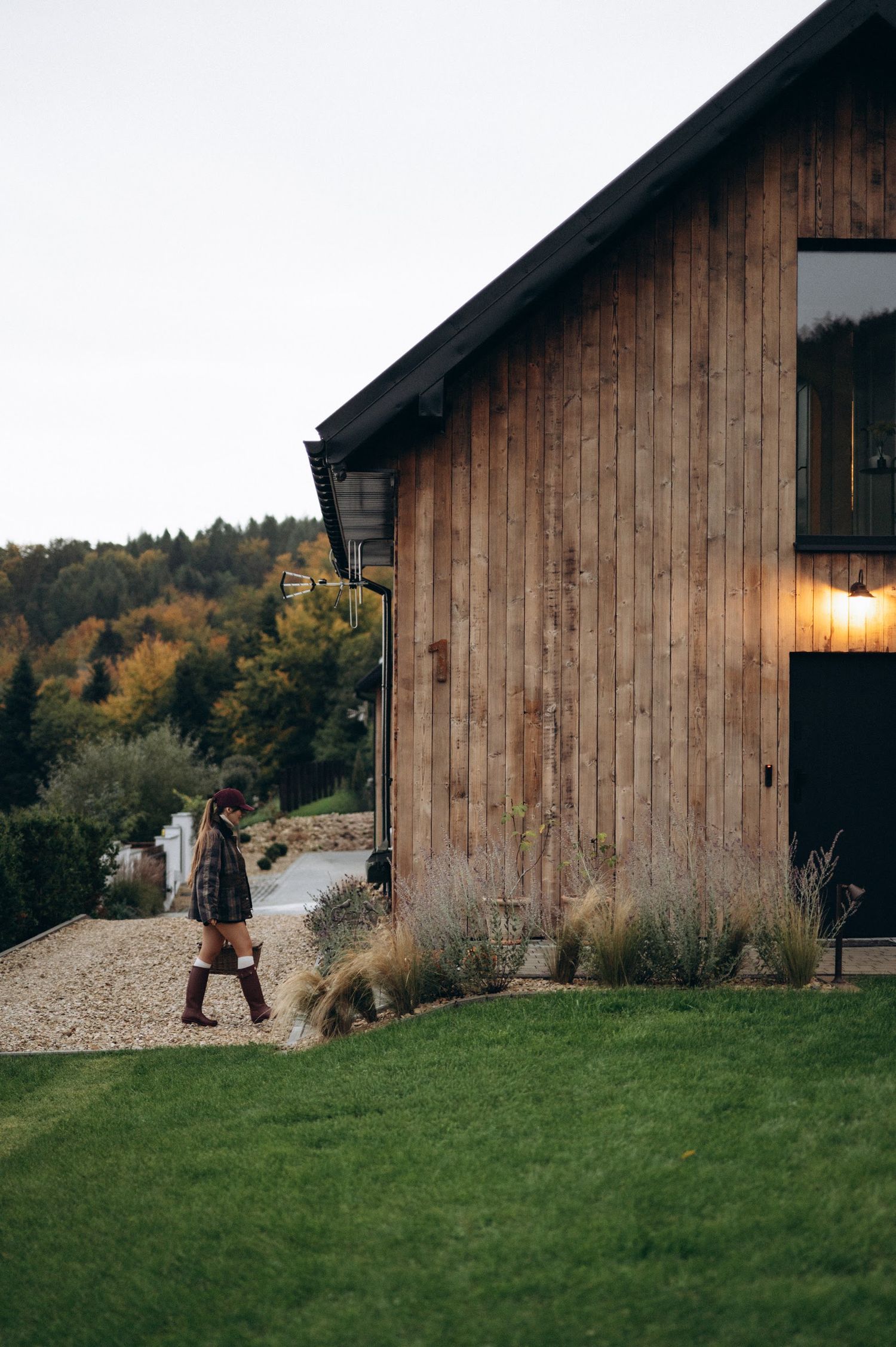 Woman walking beside a rustic wooden barn house with warm exterior lighting at dusk.