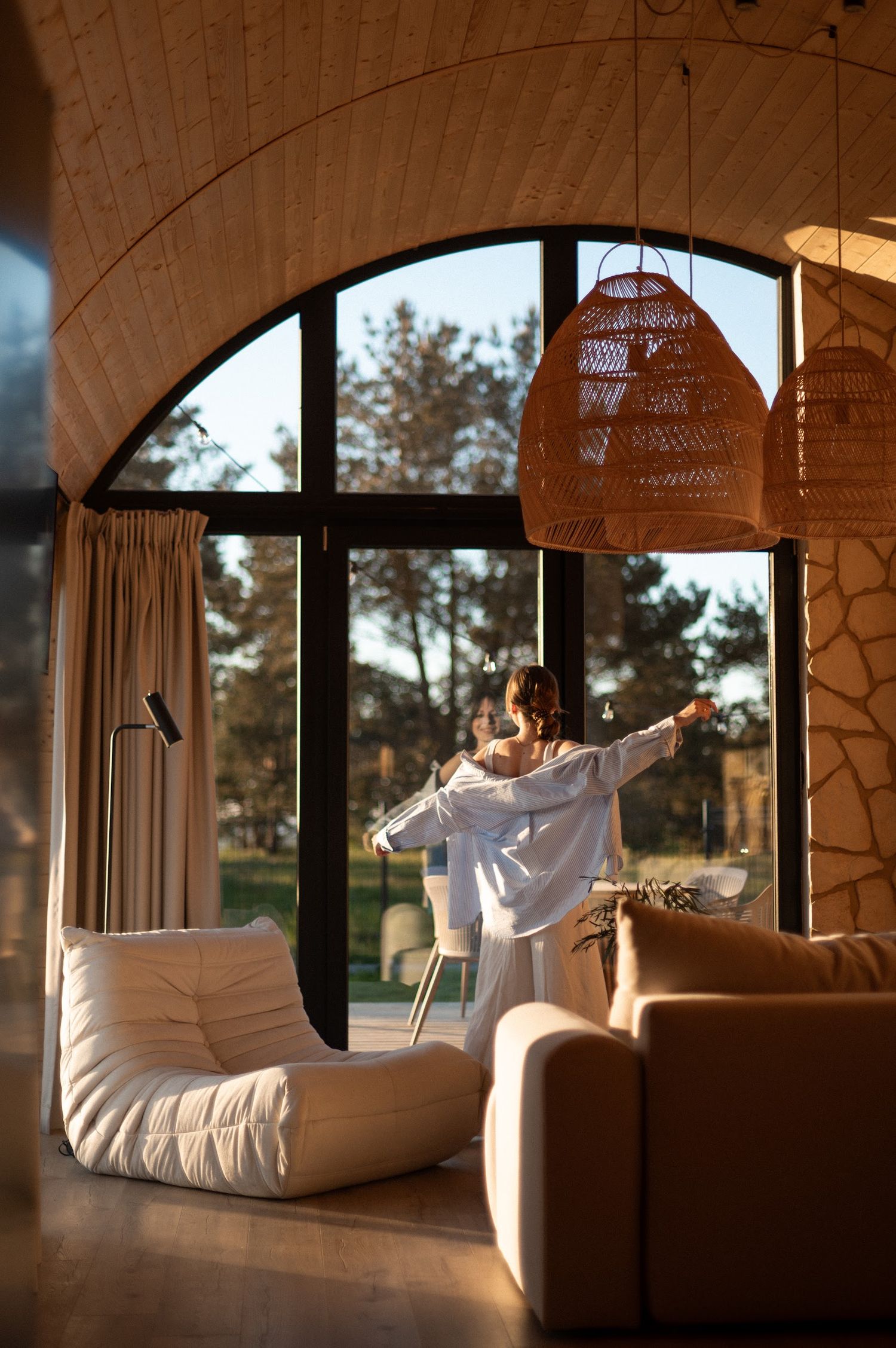 Woman standing by large arched window in warm sunlit modern interior with rattan pendant lights and cozy sofa.