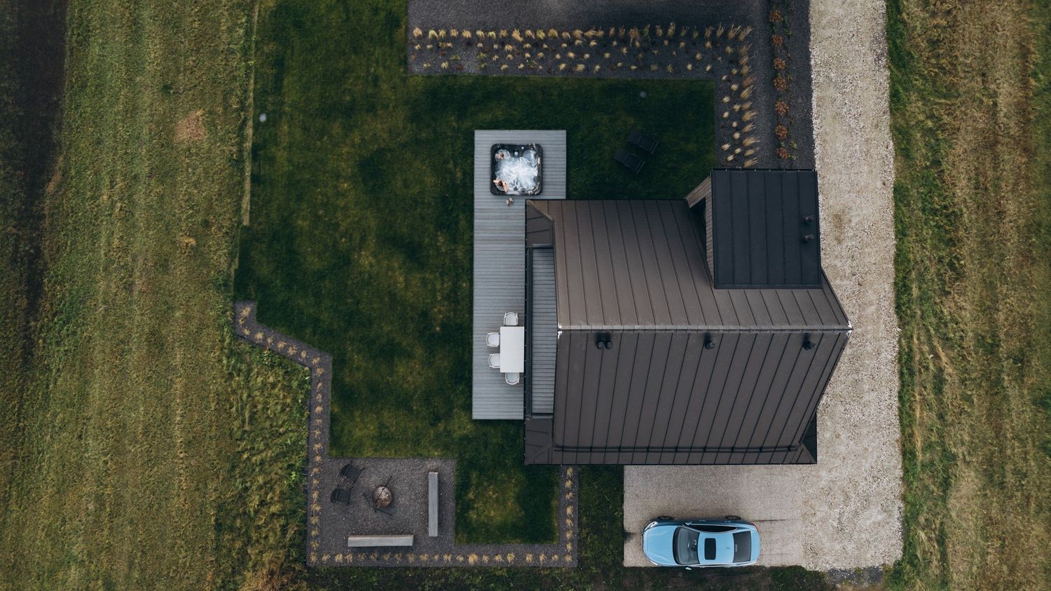 Aerial top-down view of a modern dark-roofed house with green lawn, paved driveway, and blue car parked outside.