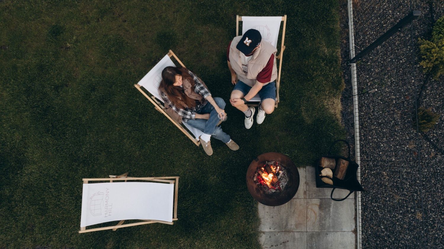 Aerial view of two people relaxing in lawn chairs around a backyard fire pit at dusk.