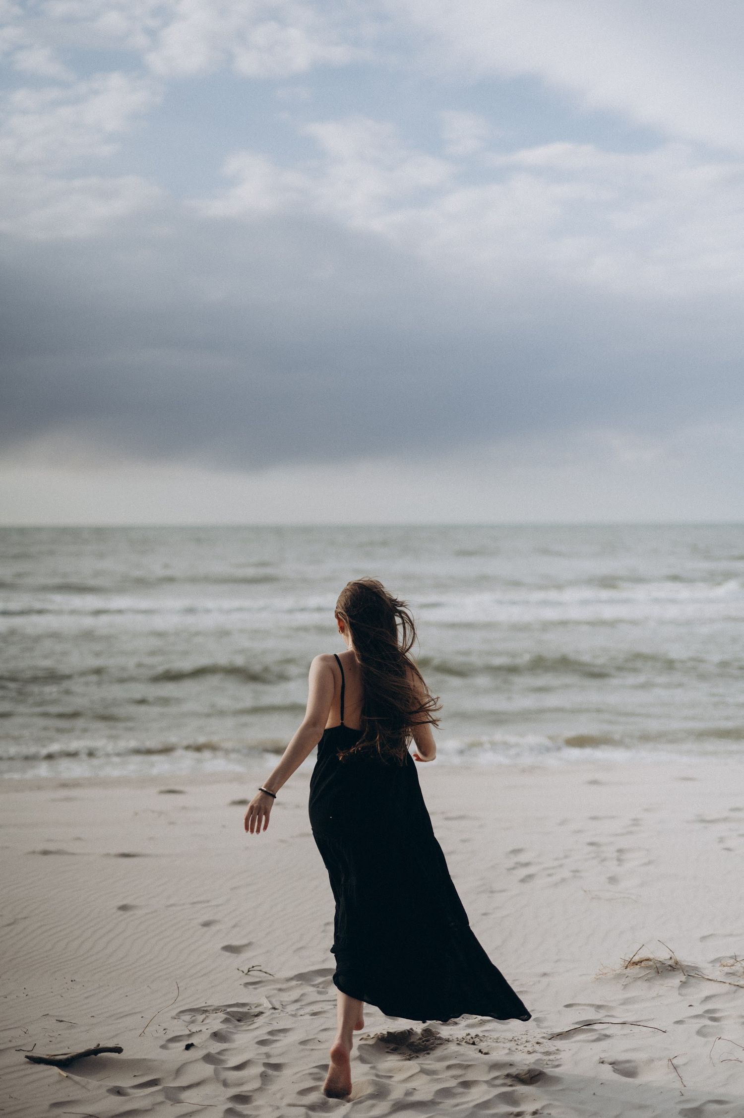 Woman in black maxi dress walking barefoot on a sandy beach toward the ocean under a cloudy sky.