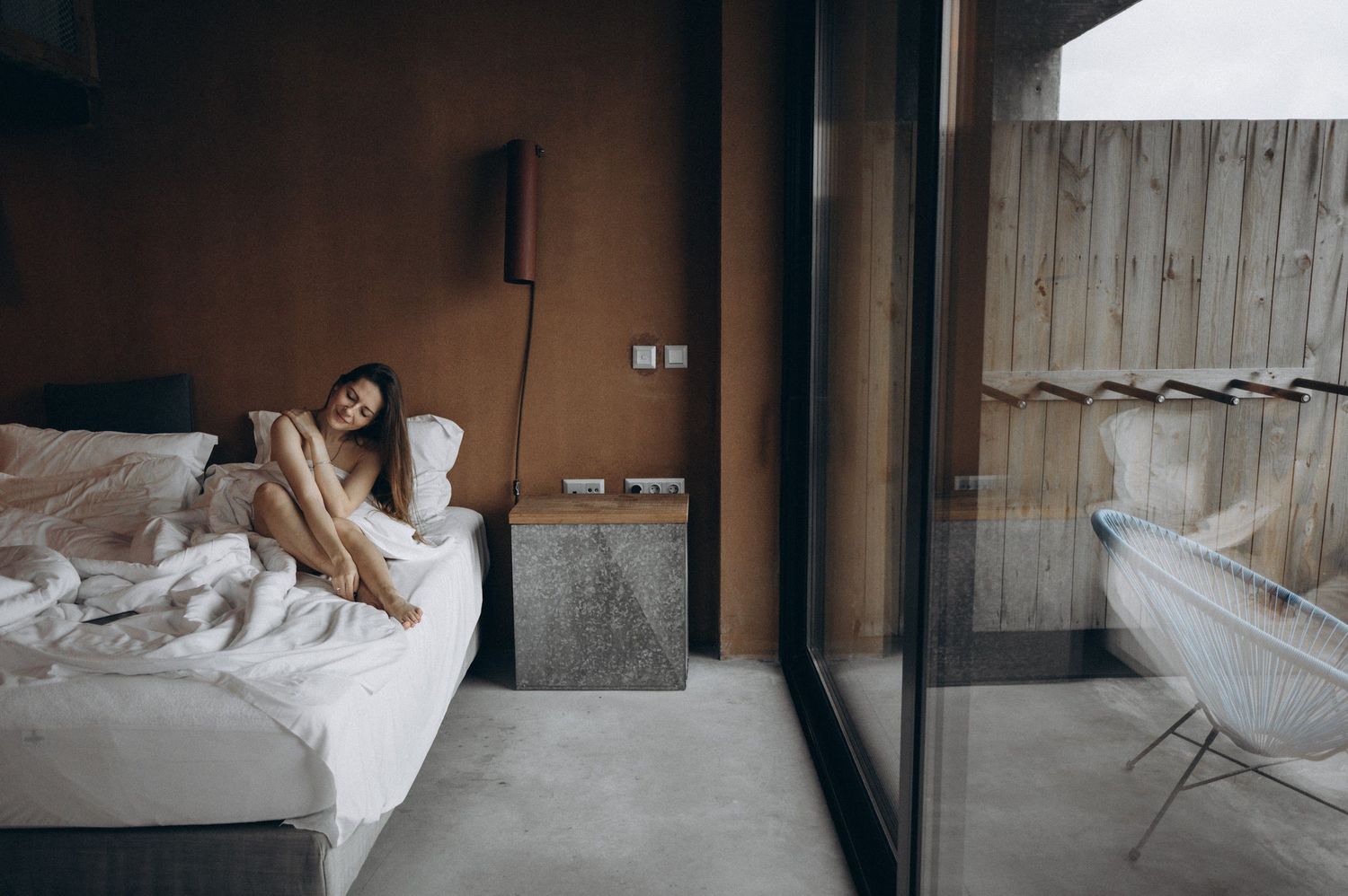 Woman sitting on a bed in a modern hotel room with large glass doors leading to a balcony with a bathtub.