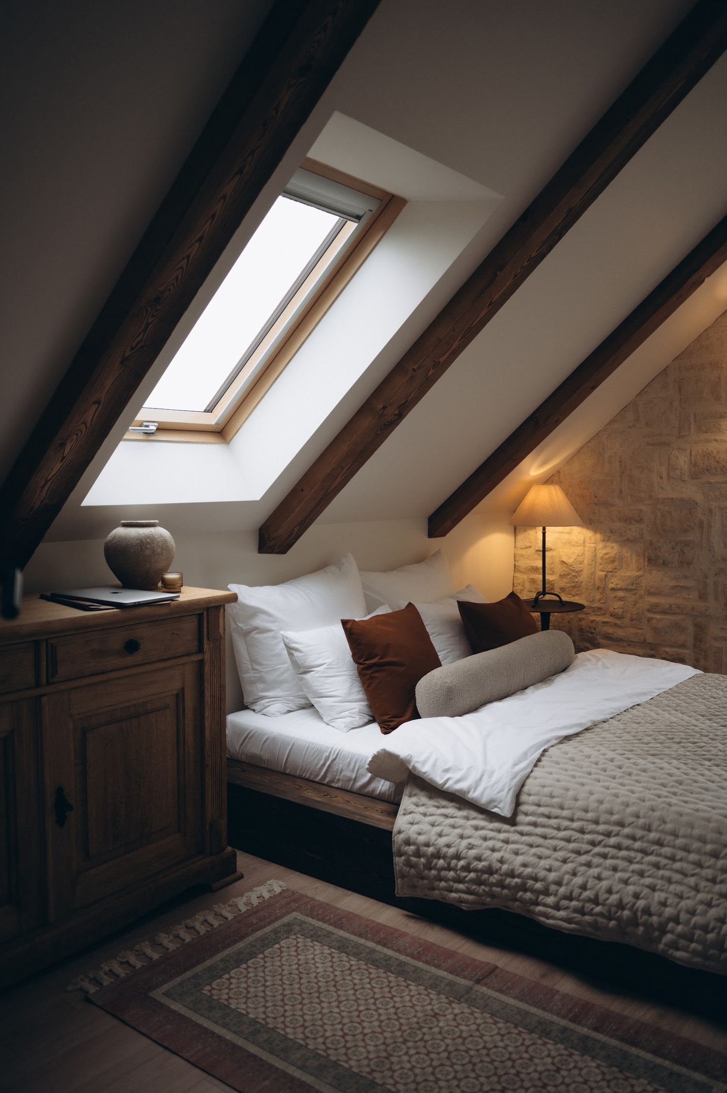 Cozy attic bedroom with exposed wooden beams, skylight window, and warm ambient lighting on a rustic bed.