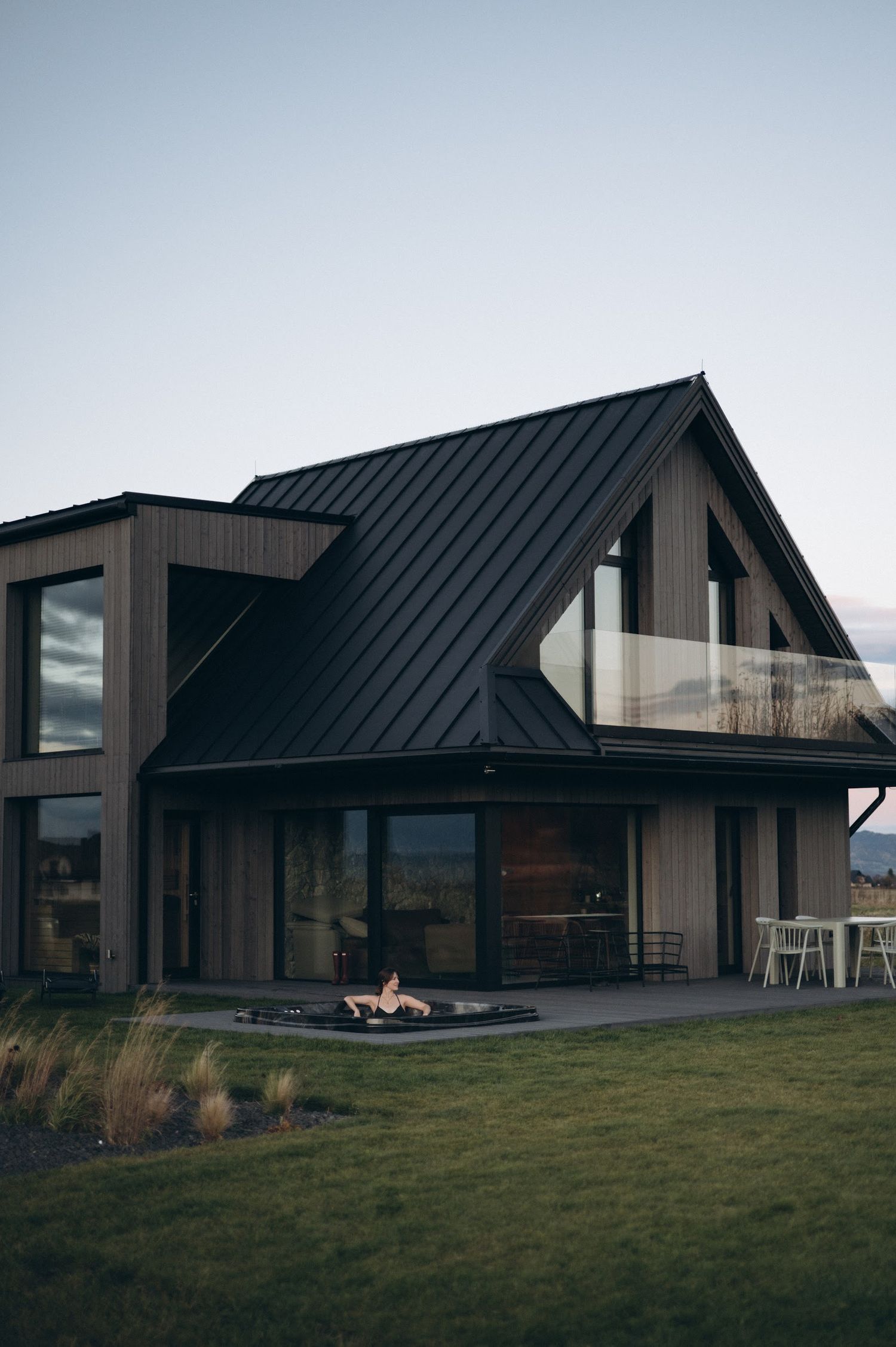 Modern dark wood and black metal roof barn-style house with large windows and fire pit at twilight.