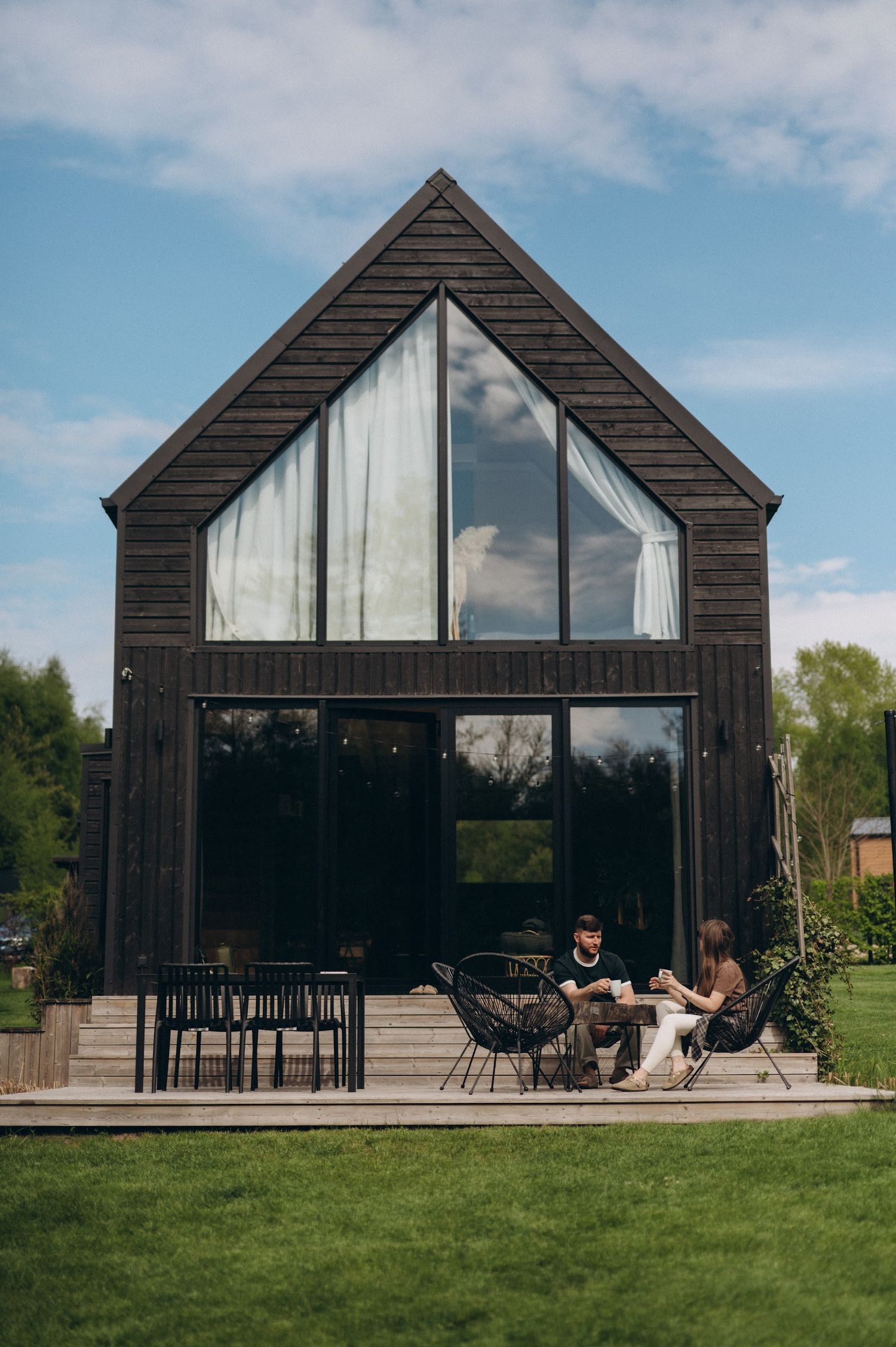 Couple relaxing on the deck of a modern dark wood barn-style house with large glass windows.