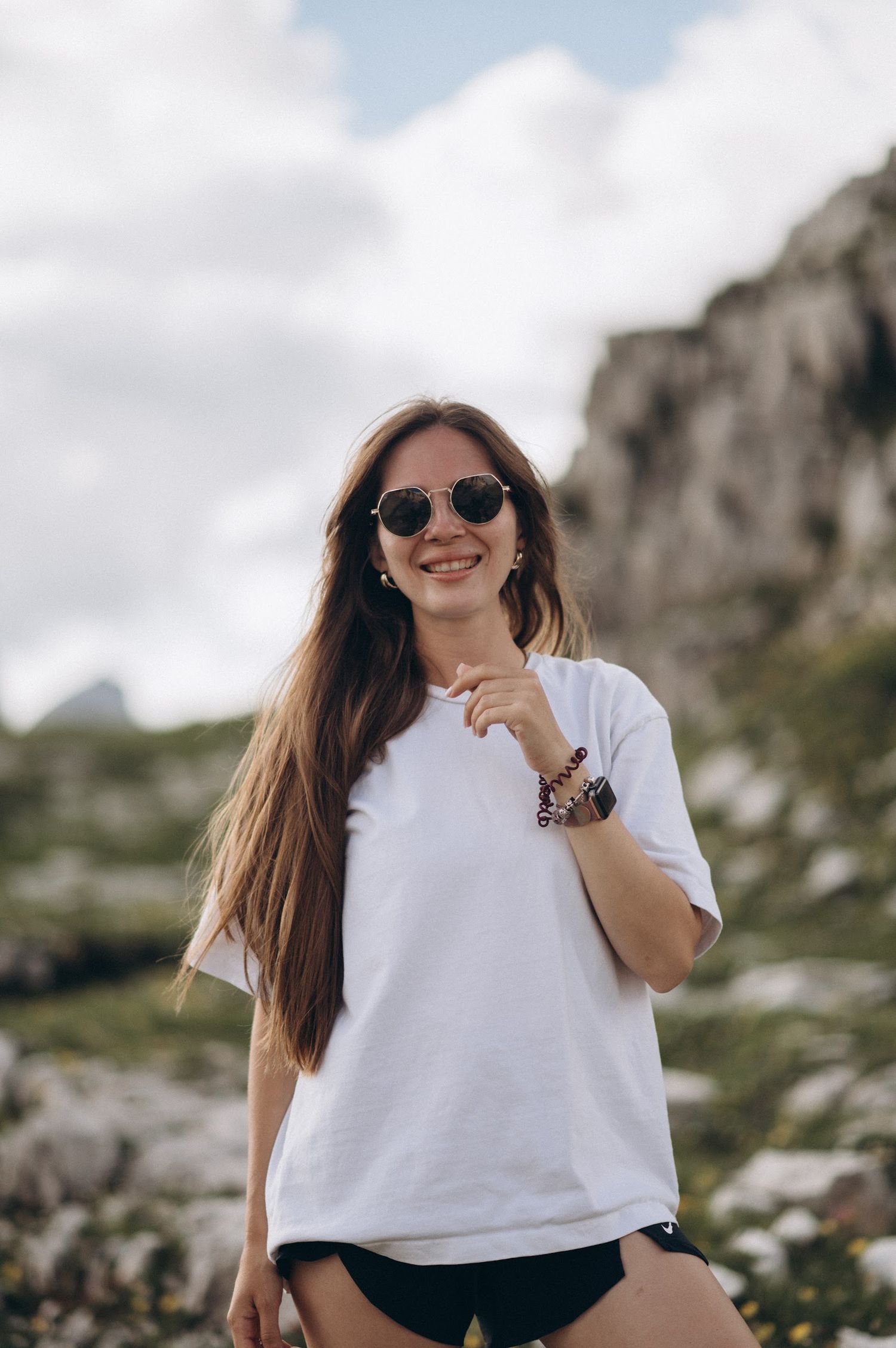 Smiling young woman in white oversized t-shirt and sunglasses posing outdoors in rocky mountain landscape.