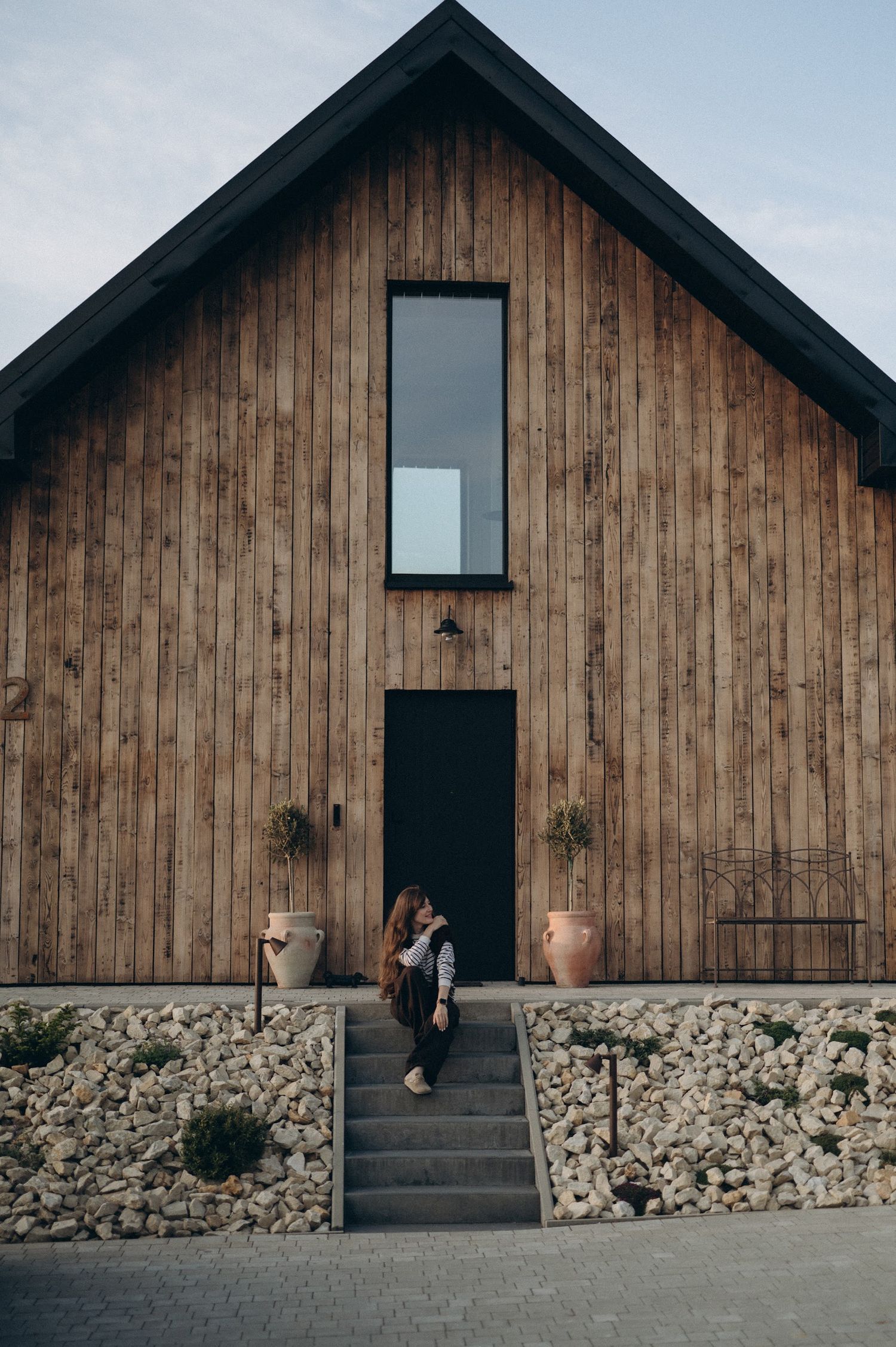 Woman sitting on stone steps at entrance of modern wooden barn house with black door and gabion stone base.