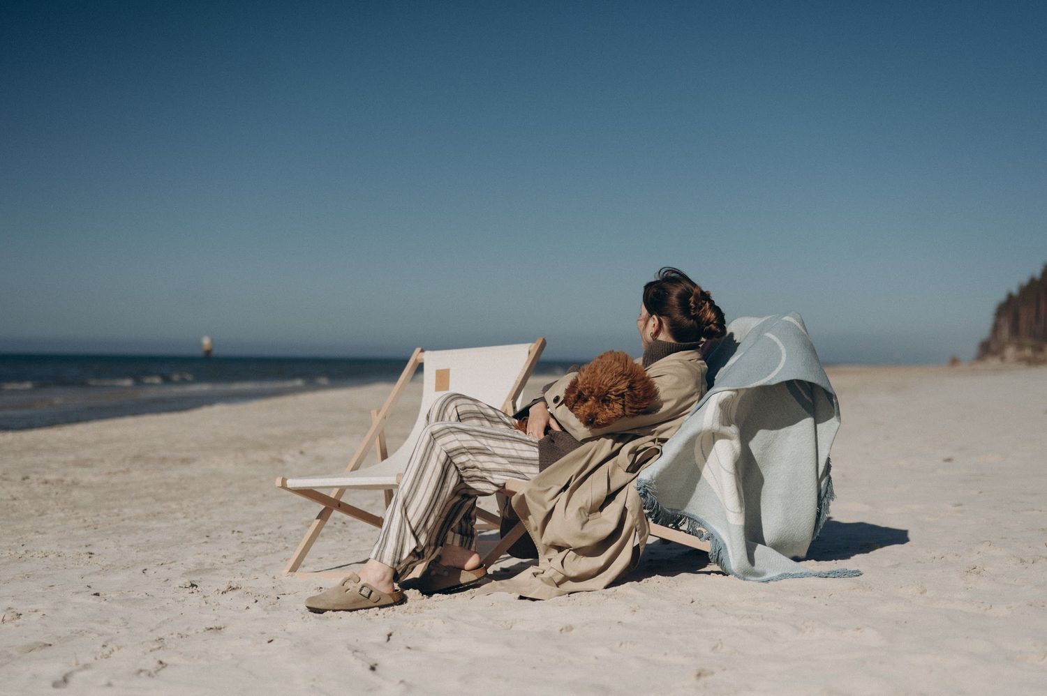 Two people relaxing in beach chairs on a sandy shore, wrapped in cozy blankets under a clear blue sky.