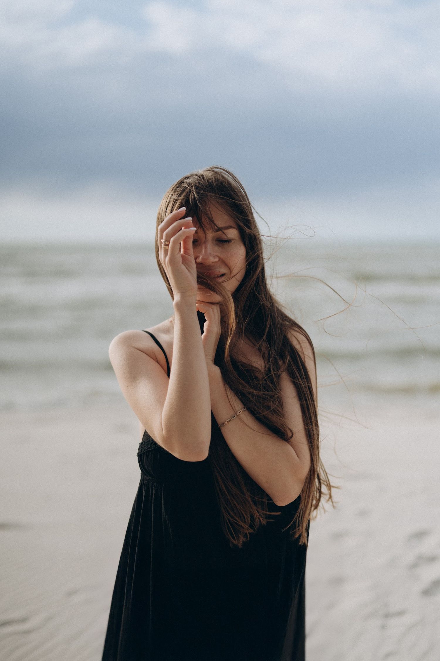Young woman in a black dress standing on a cloudy beach with wind blowing her hair.