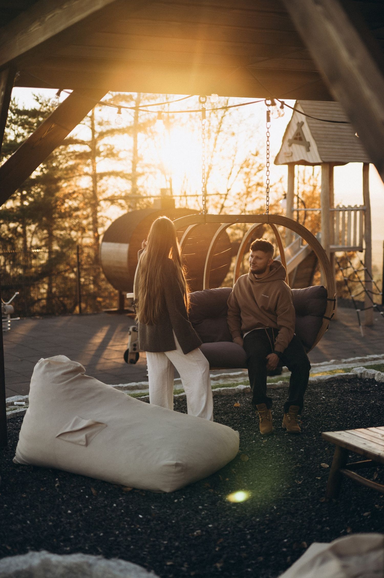 Couple relaxing at sunset on outdoor terrace with hanging egg chair, bean bag, and golden forest backdrop.