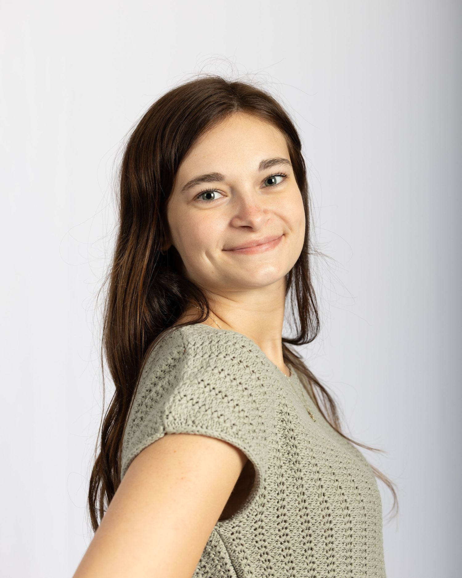 Smiling young woman with long brown hair wearing sage green knit top on white background.