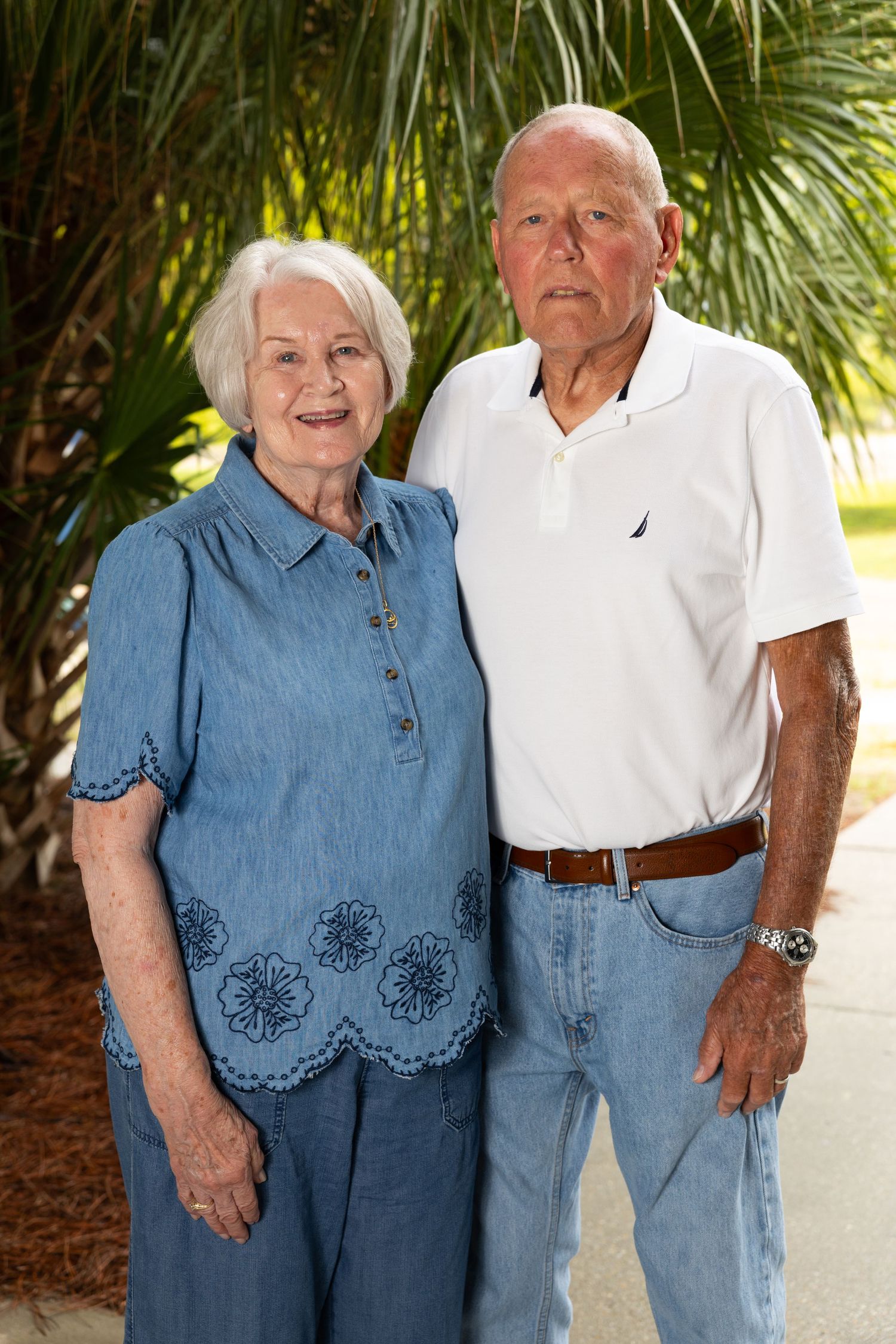 Elderly couple smiling outdoors near palm trees, woman in blue denim top and man in white polo shirt with jeans.